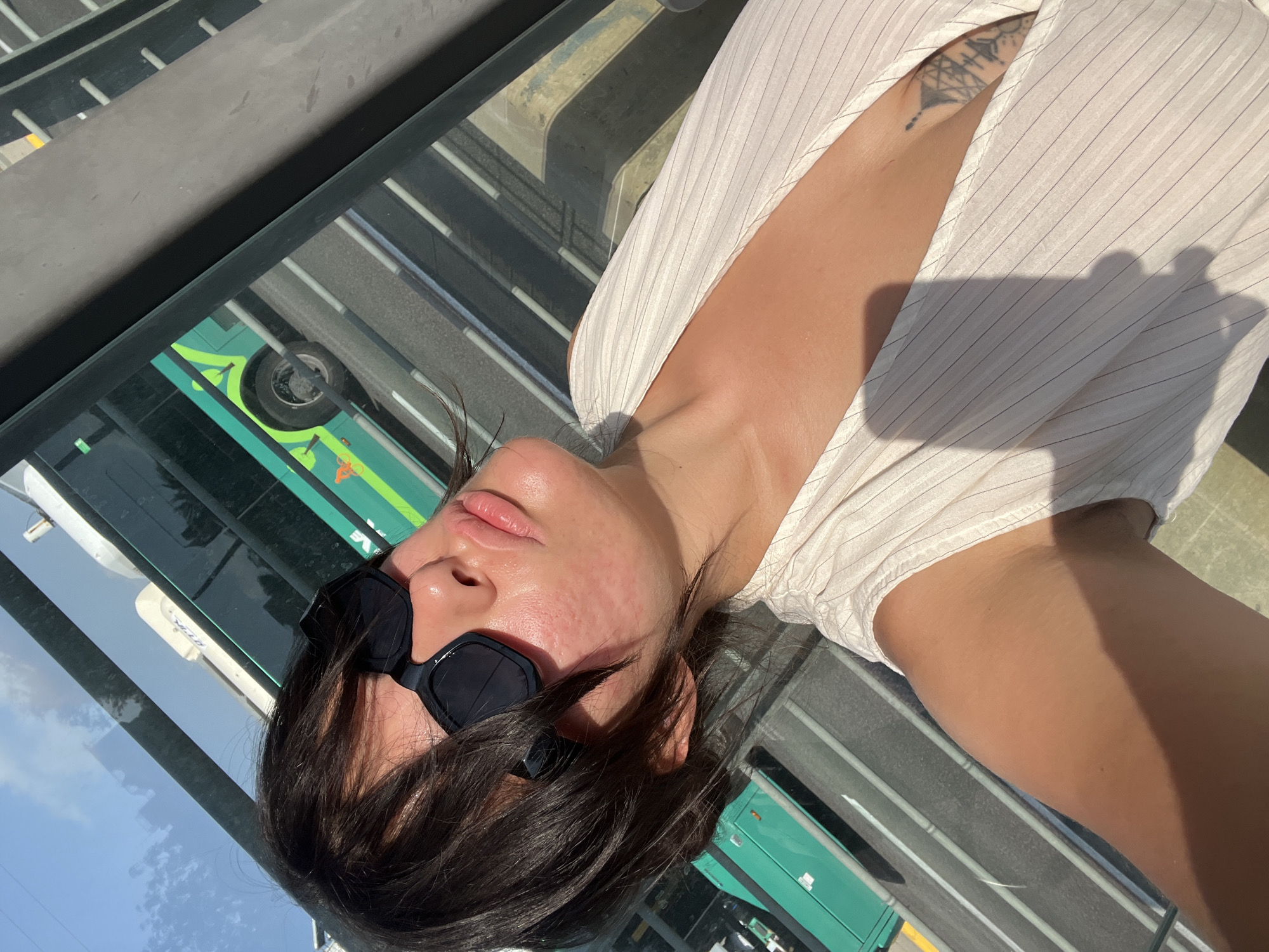 A woman with dark hair and sunglasses poses for a selfie in a city setting. She is wearing a light-colored, pinstriped top with a low neckline, revealing a tattoo on her chest. The background shows a bright sky with some clouds and a green bus on a street, suggesting an outdoor urban environment. The image appears to have been taken during the day, with sunlight casting shadows. The location is identified as Kiryat Motzkin, Israel.