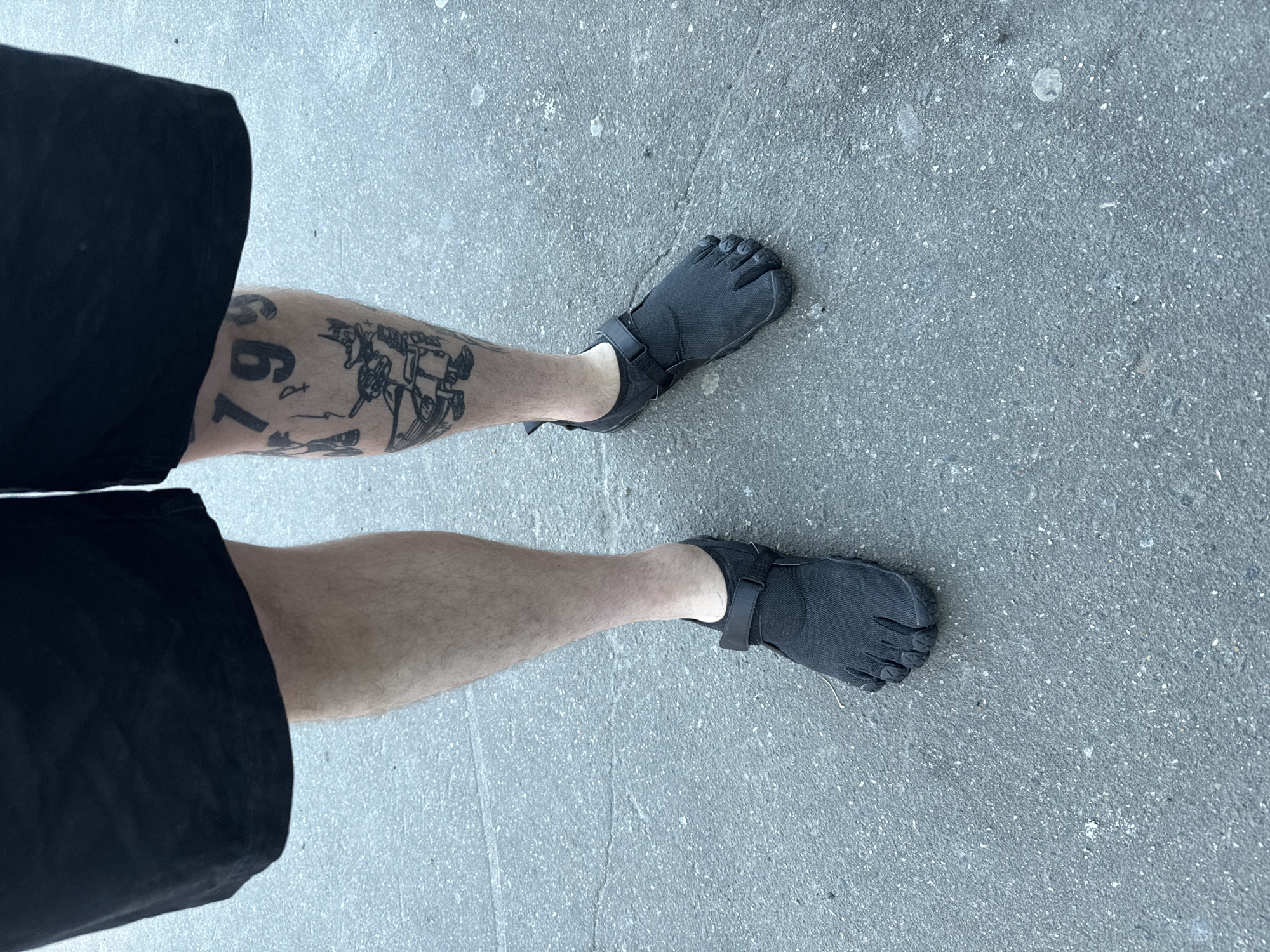 The image, taken from an overhead perspective, shows the lower half of a person's body from the waist down to the feet, standing on a paved outdoor surface in Moscow, Russia. The individual is wearing black shorts and a pair of dark gray or black minimalist 'toe shoes,' characteristic of designs like Vibram FiveFingers, which feature individual compartments for each toe and a strap over the instep.
The person's legs are visible, with the left leg adorned with several black ink tattoos. Prominent numerical tattoos include "679" and partially visible "1793". Alongside these numbers are illustrative tattoos depicting figures or scenes, the specifics of which are not entirely clear but appear to be narrative or thematic in design. The right leg is visible and appears to be untattooed.
The ground is a light gray, coarse-textured asphalt or concrete, consistent with an urban or suburban outdoor environment. The lighting is bright and even, suggesting daytime and clear weather conditions without strong shadows. The person's posture indicates they are either standing or paused while walking.