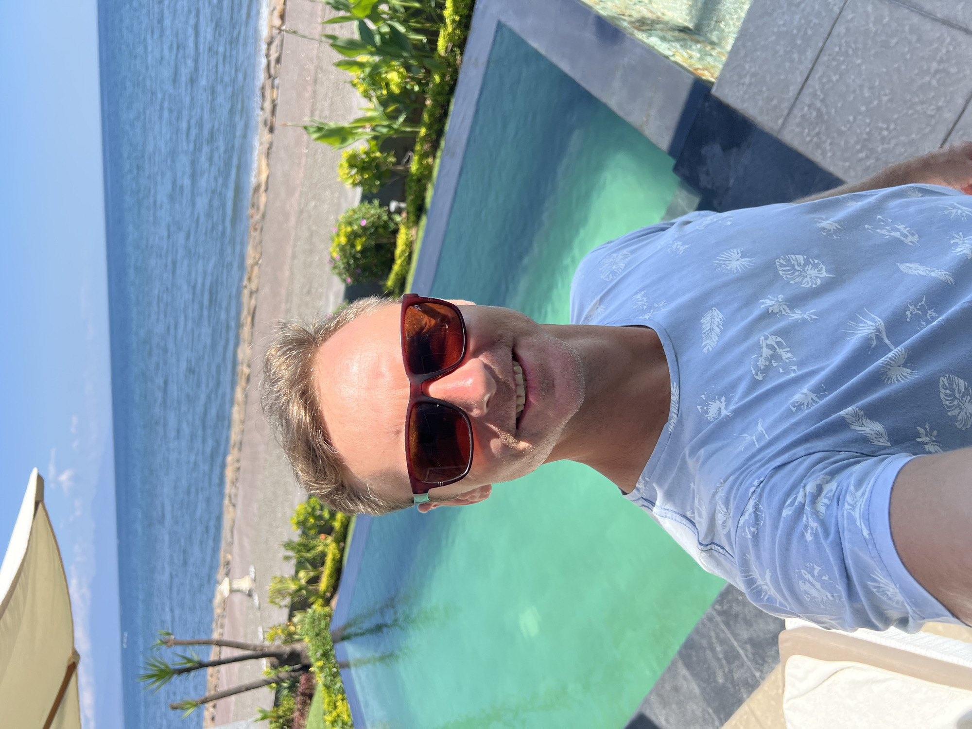 A man with blond hair and sunglasses takes a selfie by a turquoise swimming pool. He is wearing a light blue t-shirt with a white leaf pattern. The pool overlooks a beach and the ocean under a clear blue sky. In the background, there are palm trees, lush green plants, and a paved walkway. The scene is likely in a tropical resort location, possibly Buleleng, Indonesia, known for its coastal beauty and resorts.