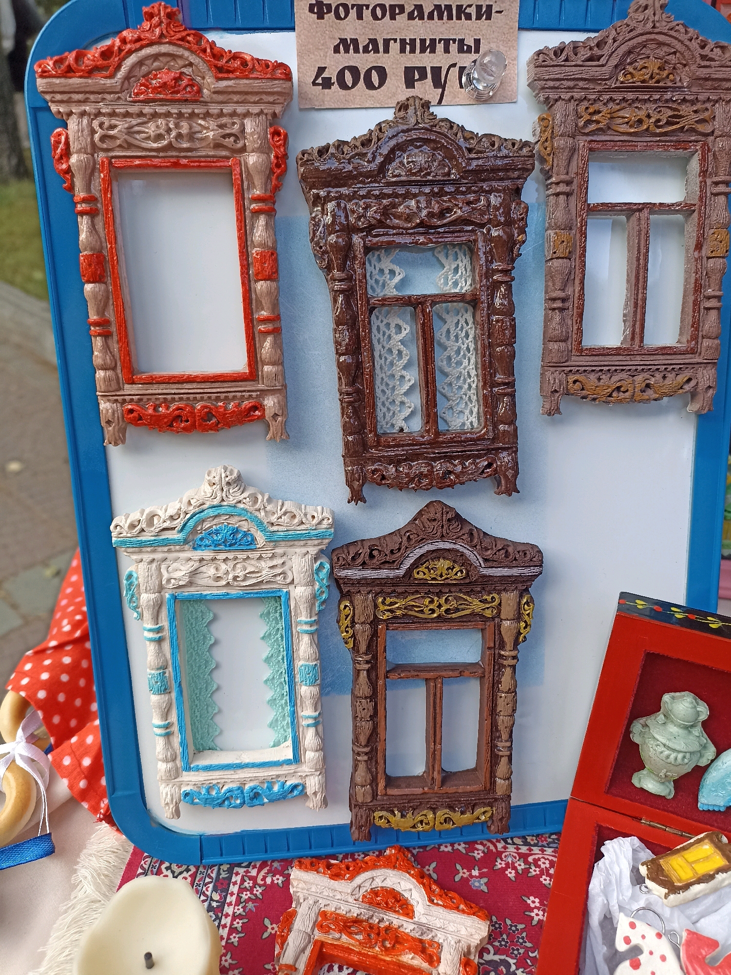 The image shows a close-up of a display of magnets shaped like traditional Russian wooden window frames, known as "nalichniki." These magnets are arranged on a white board with a blue border, which is leaning against an unseen structure. A sign above the magnets reads "ФОТОРАМКИ-МАГНИТЫ 400 РУБ," indicating that these are photo frame magnets priced at 400 rubles. The magnets themselves are intricately carved and painted in various colors, including brown, white with blue accents, and a reddish-orange. Some of the magnets have lace-like patterns visible through the "window" panes.
In the lower right corner, a red open box reveals several small decorative items, including a light green ceramic figurine of what appears to be a frog or similar creature, and another small window frame magnet with yellow paint. A white cloth with red polka dots is visible on the left side of the image, along with a bagel on a string tied with blue ribbon. A white candle is also visible in the foreground. The background is blurred but suggests an outdoor setting with greenery, consistent with a market or street fair.
The items are likely being sold at a local market or craft fair in Novosibirsk, Russia, given the Cyrillic text on the sign and the traditional nature of the crafts. No individuals are clearly visible, though the presence of a market suggests potential shoppers or vendors are nearby. The lighting indicates it is daytime.