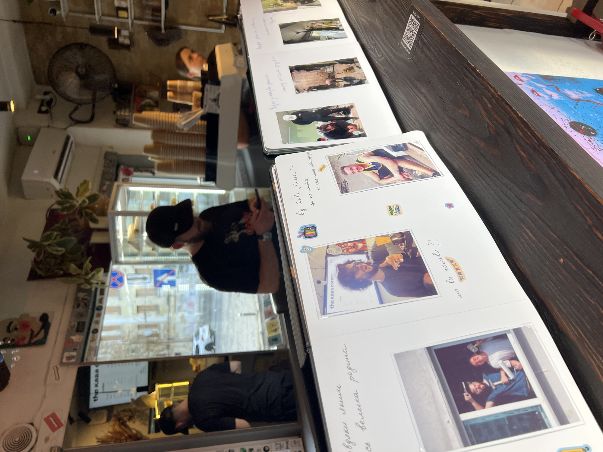 The image depicts the interior of a cafe in Kyiv, Ukraine. The perspective is at a slight downward angle, focusing on a wooden counter displaying an open photo album and several framed photographs. In the background, a window reflects the street scene, showing a street, a building, and a white car.
Inside the cafe, two men wearing black caps are visible. One man, positioned on the left and facing away from the camera, is wearing a black t-shirt. The other man, on the right, is also wearing a black t-shirt and appears to be holding a phone. A woman with dark hair is visible behind the counter on the right, looking towards the front.
The photo album open on the counter contains several photos. One photo shows a man with curly hair, wearing sunglasses and a dark shirt, standing in front of a computer screen that displays the text "the kaвa corner". Another photo on the left page shows two men holding hammers and looking out of a window. The right page of the album displays multiple smaller photos, including one of a man in a white t-shirt and a second man in dark clothing crouching, and another of people working on what appears to be construction or renovation. Some of the photographs have handwritten text in Ukrainian.
On the counter, there are stacks of paper cups and other cafe equipment. A QR code is visible on the right side of the counter. The overall ambiance suggests a casual, perhaps social, gathering or documentation of events.