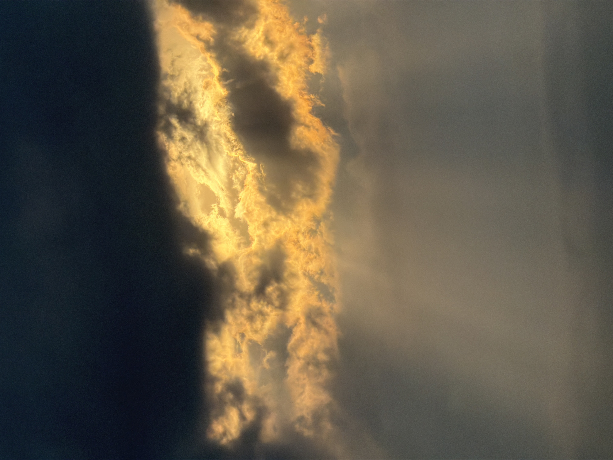 The image shows a dramatic sky. On the left side of the image, there's a dark, almost black cloud formation. In the center, a column of golden clouds is illuminated by the sun, with rays of light visible through gaps in the clouds. The right side of the image fades into a gray, hazy sky.
This image is probably a photograph of clouds during sunset or sunrise. The specific location in Tbilisi, Georgia, cannot be determined from the visual information provided.