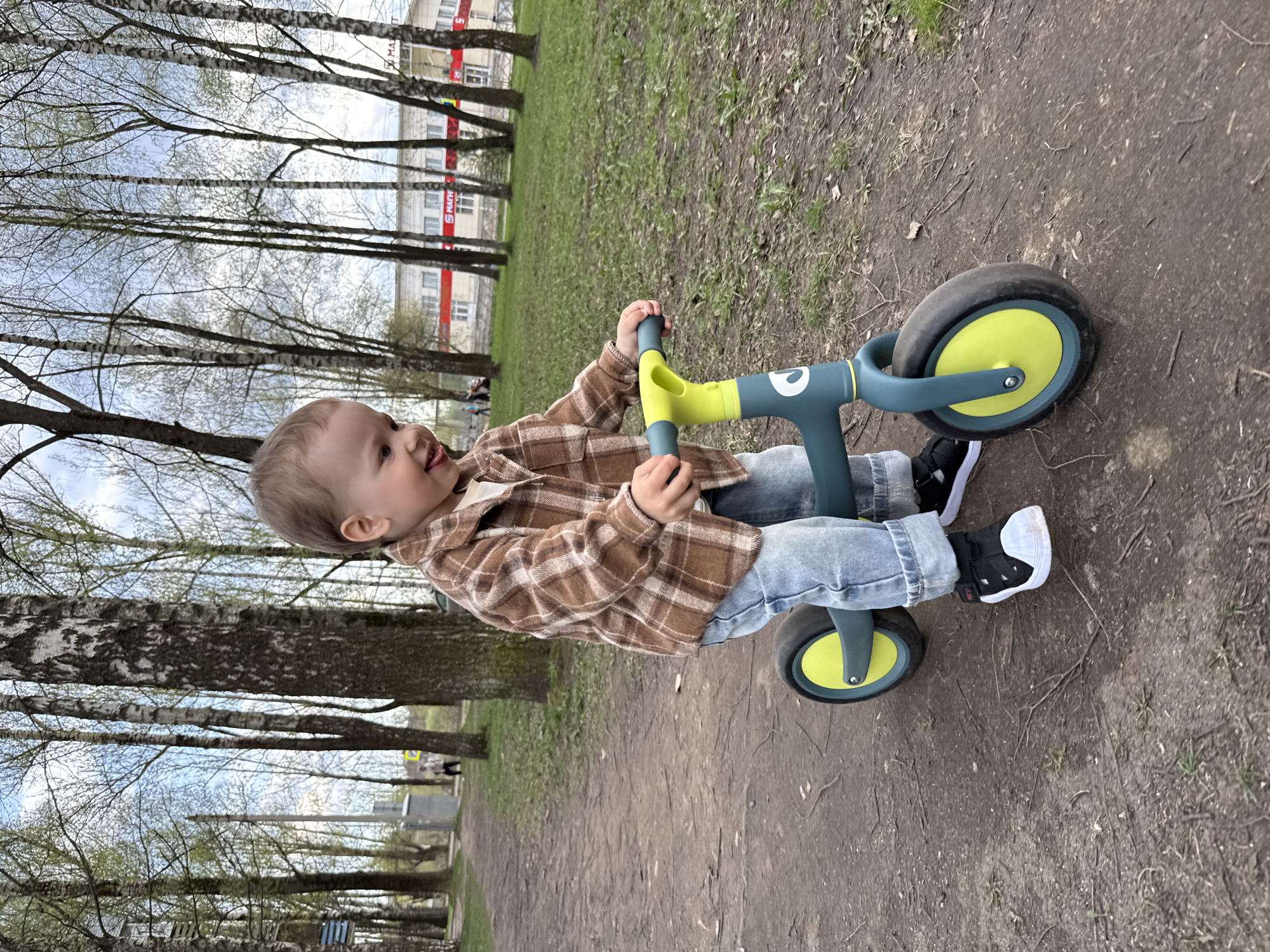 A toddler is riding a balance bike on a dirt path, with trees lining the background. The child wears a brown plaid shirt, light blue jeans, and black and white sneakers. The balance bike has a grey frame and yellow wheels. The location appears to be a park in Rusanovo, Russia, based on the signage visible on a building in the background. The surrounding environment consists of trees, grass, and a dirt path.