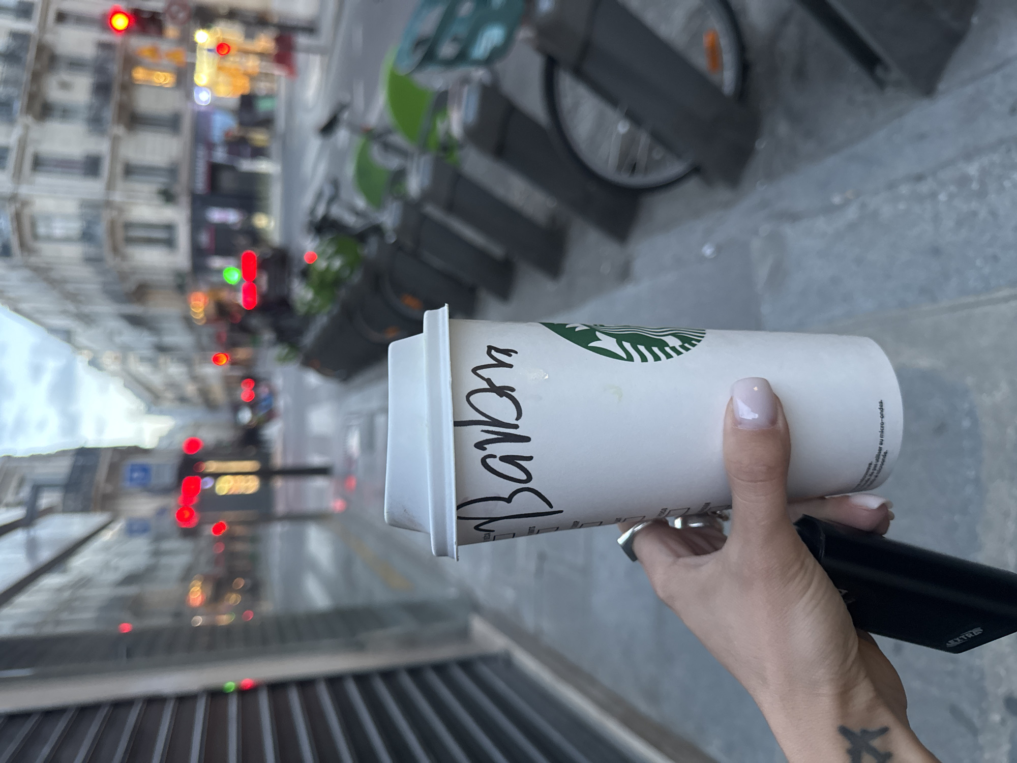 The image captures a close-up of a person's hand holding a white Starbucks coffee cup and a black rectangular electronic device, set against the backdrop of a busy urban street. The hand, which has light-colored nail polish and a silver ring on one finger, features an airplane tattoo on the wrist/forearm.
The Starbucks cup is adorned with its iconic green logo, and the name "Barba" (or "Barban") is handwritten in black marker on its side. French text, partially visible below the lid, reads "à utiliser au micro-ondes." (to be used in microwave). The black device held by the hand has the letters "XTR..." printed on its side.
In the midground, a row of green and grey public rental bicycles, consistent with Vélib' bikes found in Paris, are parked. The background reveals a street lined with multi-story buildings, typical of Parisian architecture. Numerous blurred red and green traffic lights, alongside other illuminated street and shop lights, suggest the time is either dusk or a bright, overcast day. The scene depicts a moment of urban life in Paris, France.
