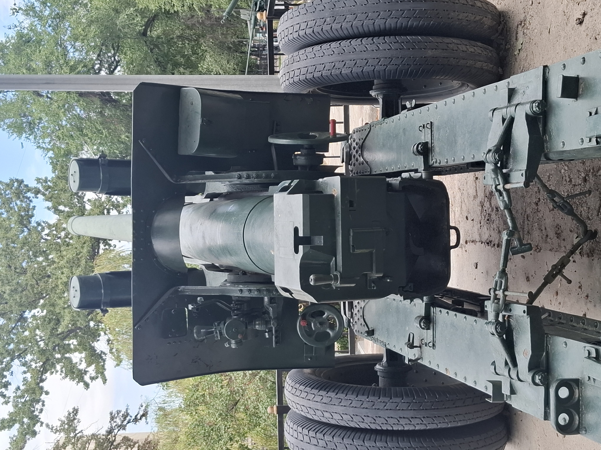 The image shows the rear view of a large, dark green artillery piece. It is mounted on a wheeled carriage with four tires in total, two on each side. The artillery piece has a prominent barrel, a complex firing mechanism with various levers and wheels, and two cylindrical protrusions on top. The object is situated outdoors, with trees and foliage visible in the background, and a light-colored pole to the right. The ground appears to be paved with some loose gravel. This artillery piece is on display at the Museum of Folk Musical Instruments named after Ykhlas in Almaty, Kazakhstan.