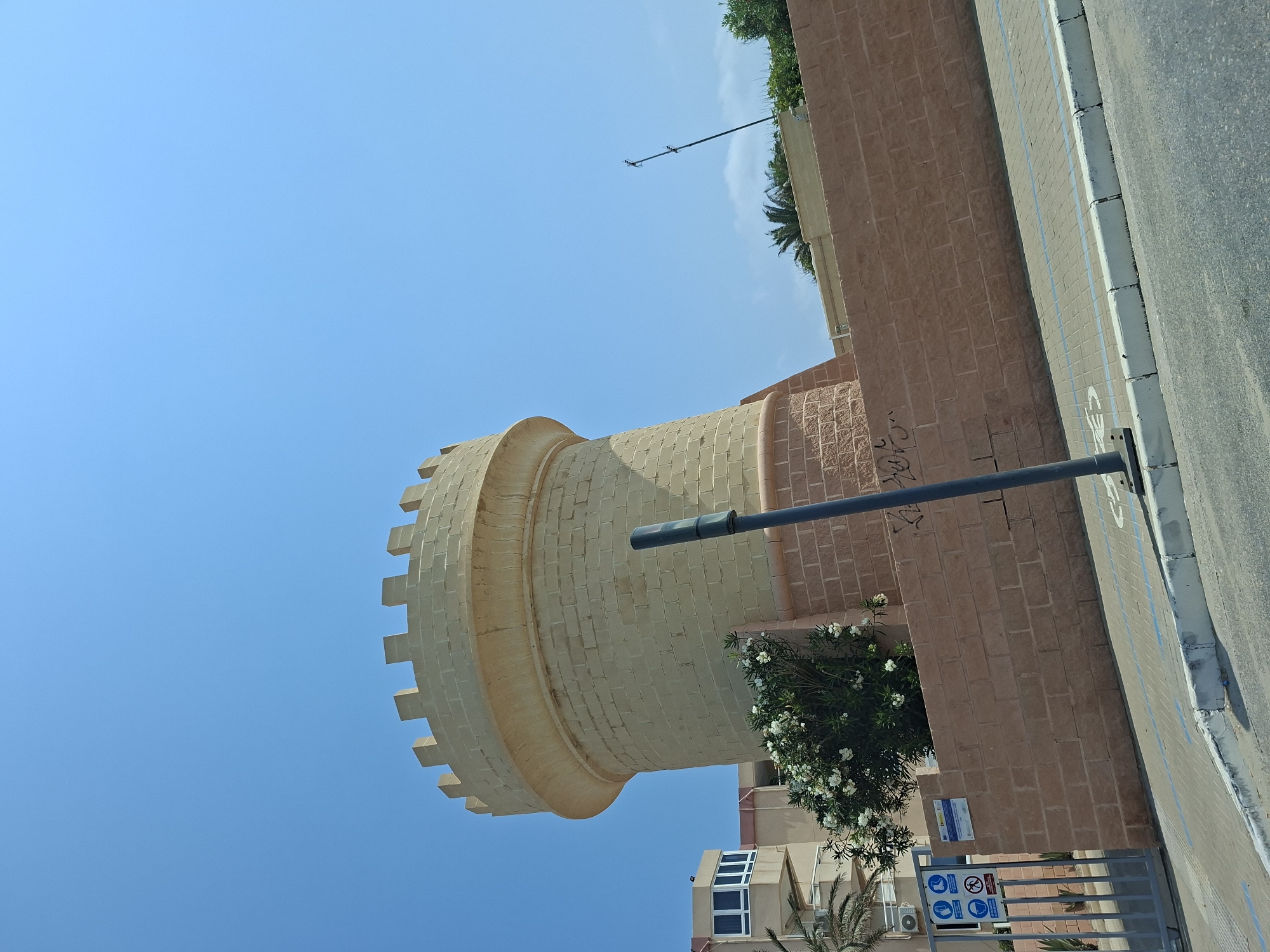 This media file captures a close-up, low-angle view of a cylindrical, stone tower with a crenellated top. The tower is constructed from pale, rectangular stone blocks. To the right of the tower is a reddish-brown brick wall, which extends upwards and to the right. A dark, horizontal pipe or pole runs across the image, partially obscuring the tower and the brick wall.
Below the tower and behind the brick wall, a modern building with multiple windows is visible, along with a lush green bush with white flowers. The foreground shows a paved walkway with blue lines, bordered by a curb and an asphalt road. Several signs are affixed to a metal fence at the bottom right, including what appears to be a safety notice with icons.
The sky is a clear, bright blue, suggesting a sunny day. The lighting indicates it is daytime, likely morning or afternoon. The scene is set in an urban or semi-urban environment, possibly a public area or a courtyard, given the walkway, road, and signage. The architectural style of the tower hints at a historical or decorative function, while the surrounding modern elements suggest a contemporary setting. No people are visible in the frame. The overall impression is one of a bright, clear day in a location with a mix of historical and modern architecture. Visible text includes signage with icons and possibly some graffiti on the brick wall. The location is identified as San Javier, Spain.