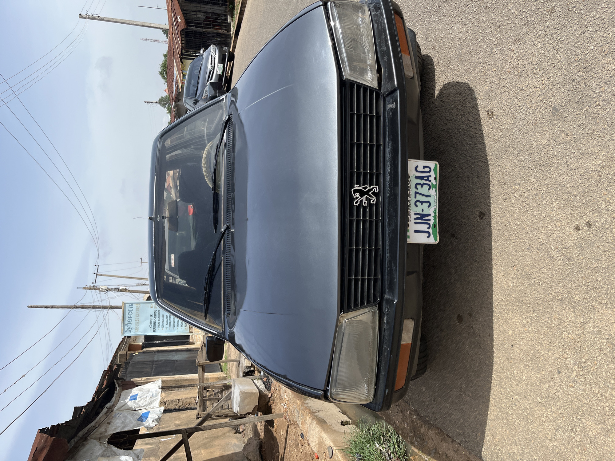 A gray Peugeot sedan is parked on the side of a road. The car's license plate reads "JJN-373AG". In the background, a building is seen with a sign that reads "Cybercom BUSINESS CENTRE" and lists services such as "COMPUTER TRAINING SCHOOL", "ONLINE REGISTRATION", "ICT CONSULTANT", "ACCESSORIES", "TYPING / PRINTING", "LAMINATION", and "BINDING". Power lines are visible overhead. The location is suggested to be Jos, Nigeria, based on the information on the license plate.