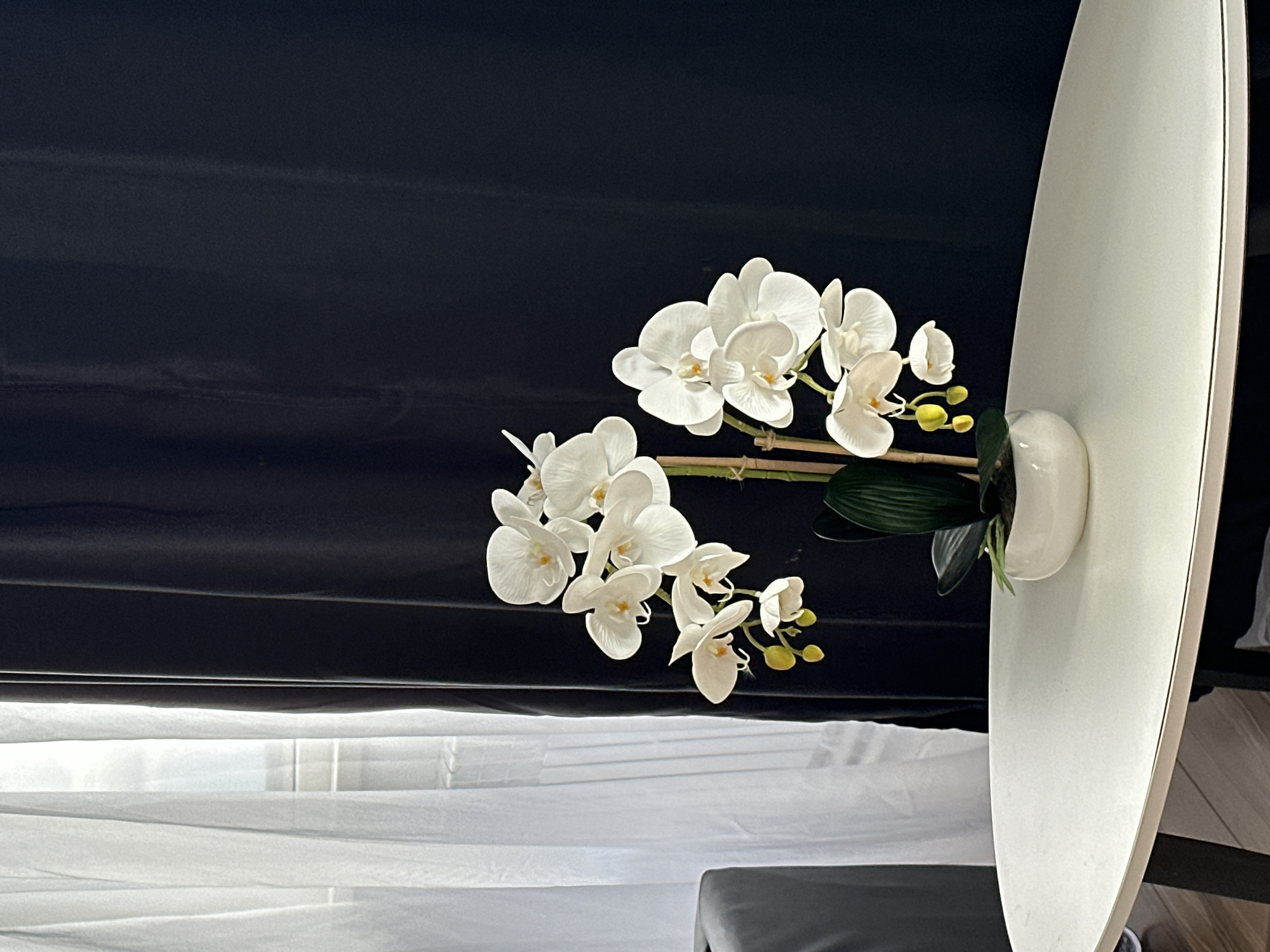 The image presents an indoor scene showcasing white phalaenopsis orchids in a white ceramic vase. The vibrant white orchid flowers, with their subtle yellow centers and green leaves, are supported by thin, light-colored stakes tied with twine. They are prominently featured against a dark, possibly black, fabric background, which appears to be a curtain or backdrop, exhibiting slight folds and texture.
On the right side of the frame, a large, smooth, light-colored object, possibly a minimalist mirror or a piece of modern furniture with a curved edge, stands vertically, partially obscuring the background. A thin, dark line runs along the edge of this object. The lower portion of the image reveals light-colored, sheer curtains or blinds, suggesting a window and filtering natural light into the space. A dark, possibly upholstered, object is partially visible in the bottom right corner.
No specific activity is taking place; the image is a still life focused on decorative elements. The lighting is soft and ambient, indicative of an indoor setting, without strong shadows to pinpoint a time of day. There is no visible text. The overall aesthetic is clean, modern, and serene. The location context provided is Chaikovsky, Russia, though no explicit visual cues within the image confirm this.