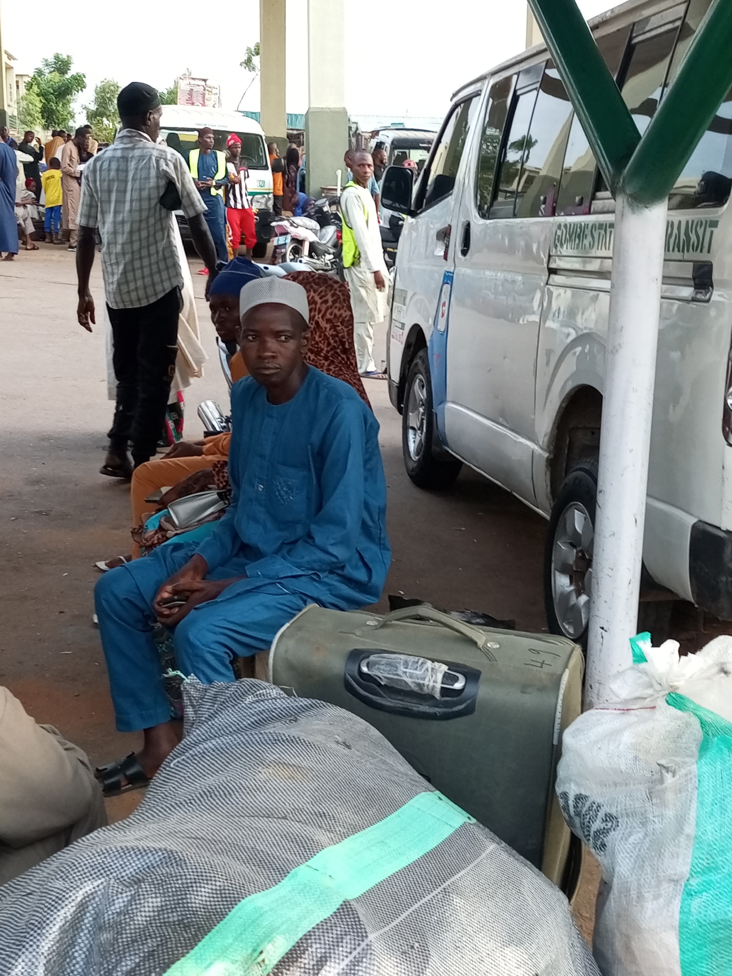 In Gombe City, Nigeria, a group of people are gathered in what appears to be a transportation hub or bus station, indicated by the presence of a "GOMBE STATE TRANSIT" bus. The scene is outdoors, with a paved ground surface. A young man, dressed in a blue traditional tunic and trousers, is seated prominently in the foreground, looking towards the camera. Beside him are luggage items, including a large, olive-green suitcase with a number "49" visible and a grey and white striped duffel bag. Other individuals are visible in the background, some standing and others seated. They are dressed in various attire, suggesting a diverse group of travelers. A white minibus is parked nearby. The overall atmosphere suggests a moment of waiting or transit.
