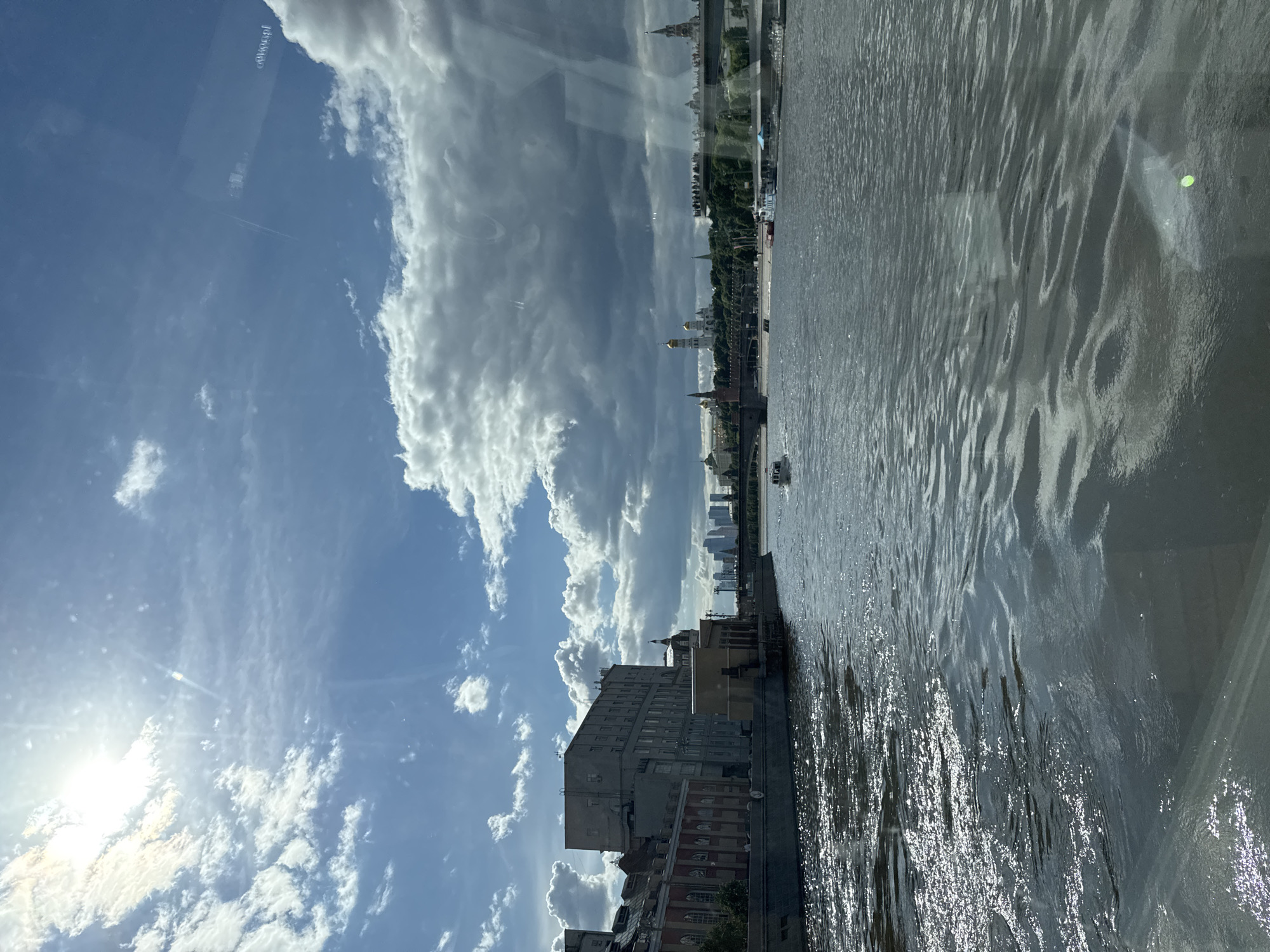 The image captures a view of the Moscow River in Russia, with the city skyline visible in the background. The river's surface reflects sunlight, creating a shimmering effect. Several buildings line the riverbank, including a large, multi-story building with a gray facade. In the distance, landmarks such as the gold domes of a cathedral and the spires of the Kremlin are visible. The sky is filled with white clouds against a blue backdrop. The sun is shining brightly, casting a glare in the lower left corner of the image. The photo appears to have been taken through glass, as there are some reflections visible.