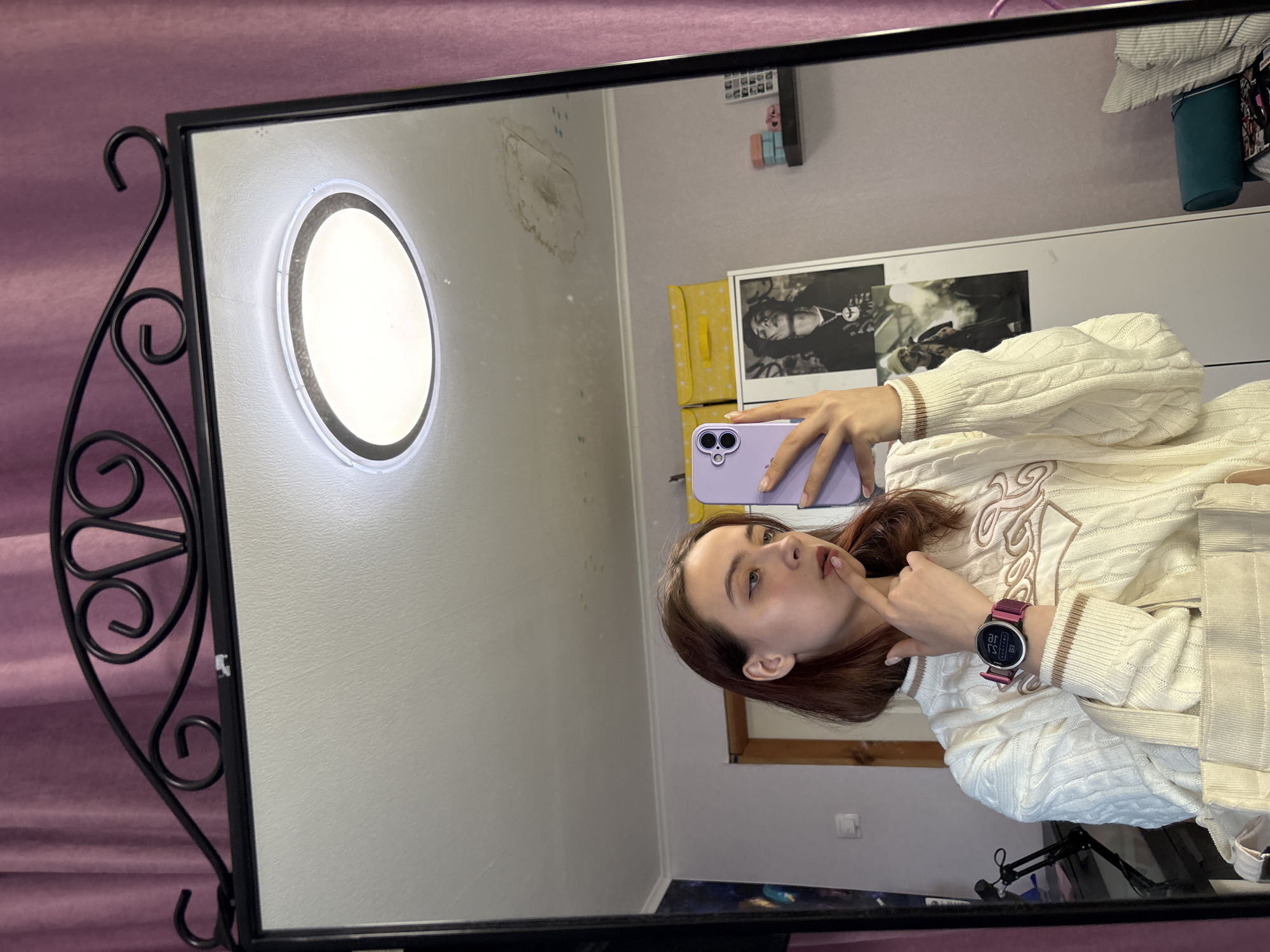 Here is a description of the image:
The image is a mirror selfie of a young woman, reflected in a decorative mirror with a black frame and ornate wrought iron detailing on the side. The mirror hangs against a pink/purple backdrop, possibly a curtain or wall covering.
The woman is holding a purple smartphone, presumably taking the selfie. She is wearing a white sweater with a decorative design, and a smartwatch with a dark face and purple band on her left wrist. Her dark hair is shoulder-length. She is touching her chin with her finger.
The reflection in the mirror shows a room with light-colored walls, a round light fixture on the wall, a framed picture above a yellow storage unit, and other items suggesting a living space or bedroom. A blue storage container is on the shelf. The location could be deduced from the interior design. The city is not specified in the image context.