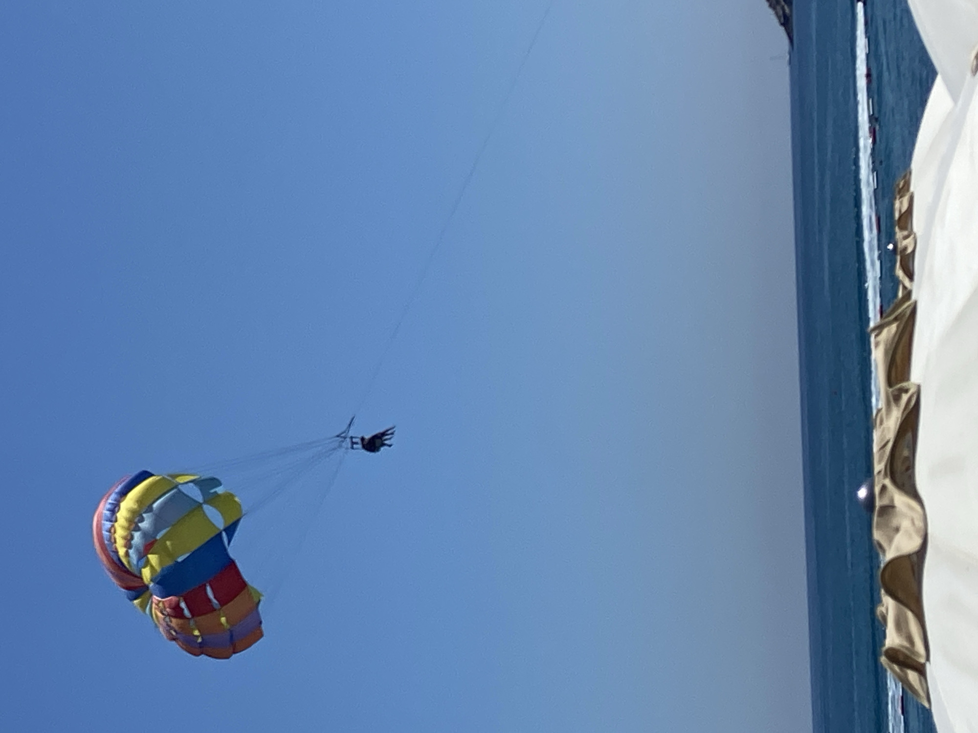 In the sky above the sea, a colorful parachute with red, orange, yellow, blue, and white segments is ascending. Two dark figures are suspended below the parachute by harnesses and lines. The parachute is positioned in the upper left quadrant of the frame. The sea, a deep blue, stretches across the lower half of the image, meeting a clear blue sky. In the foreground, the tops of two white beach umbrellas are visible, with one having a tan-colored fabric canopy partially obscuring it. The scene suggests a coastal resort area. The location is identified as Prijevor, Montenegro.