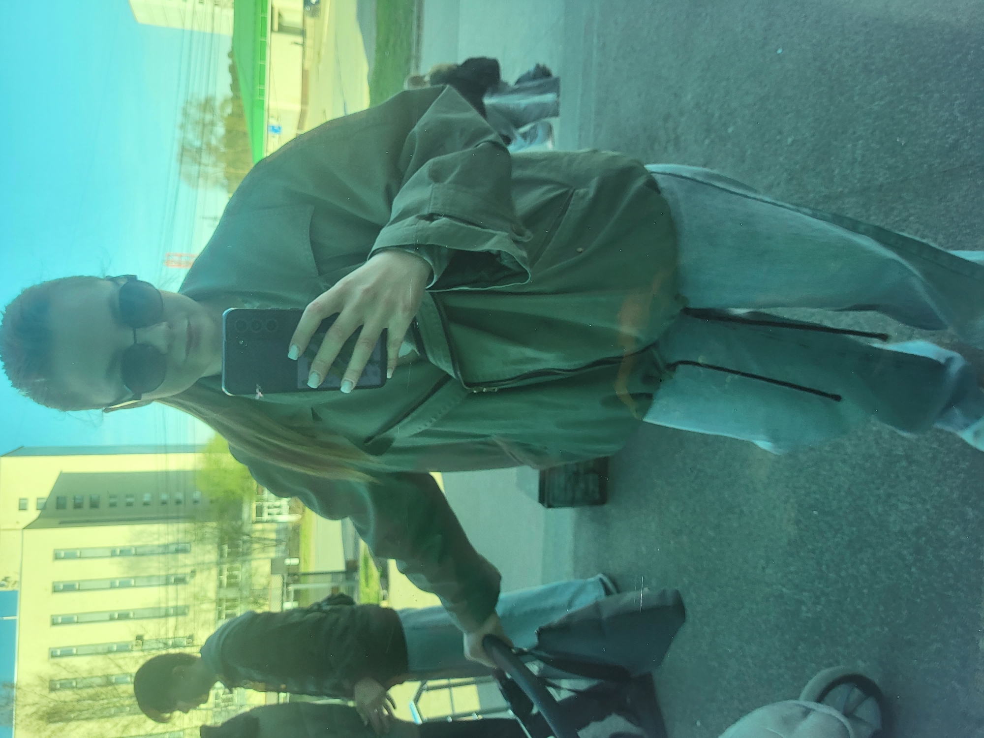 A woman is taking a selfie. She is wearing sunglasses, a green jacket, and jeans. The reflection in the window shows a building with many windows. A person is pushing a stroller. The location is Chaikovsky, Russia.