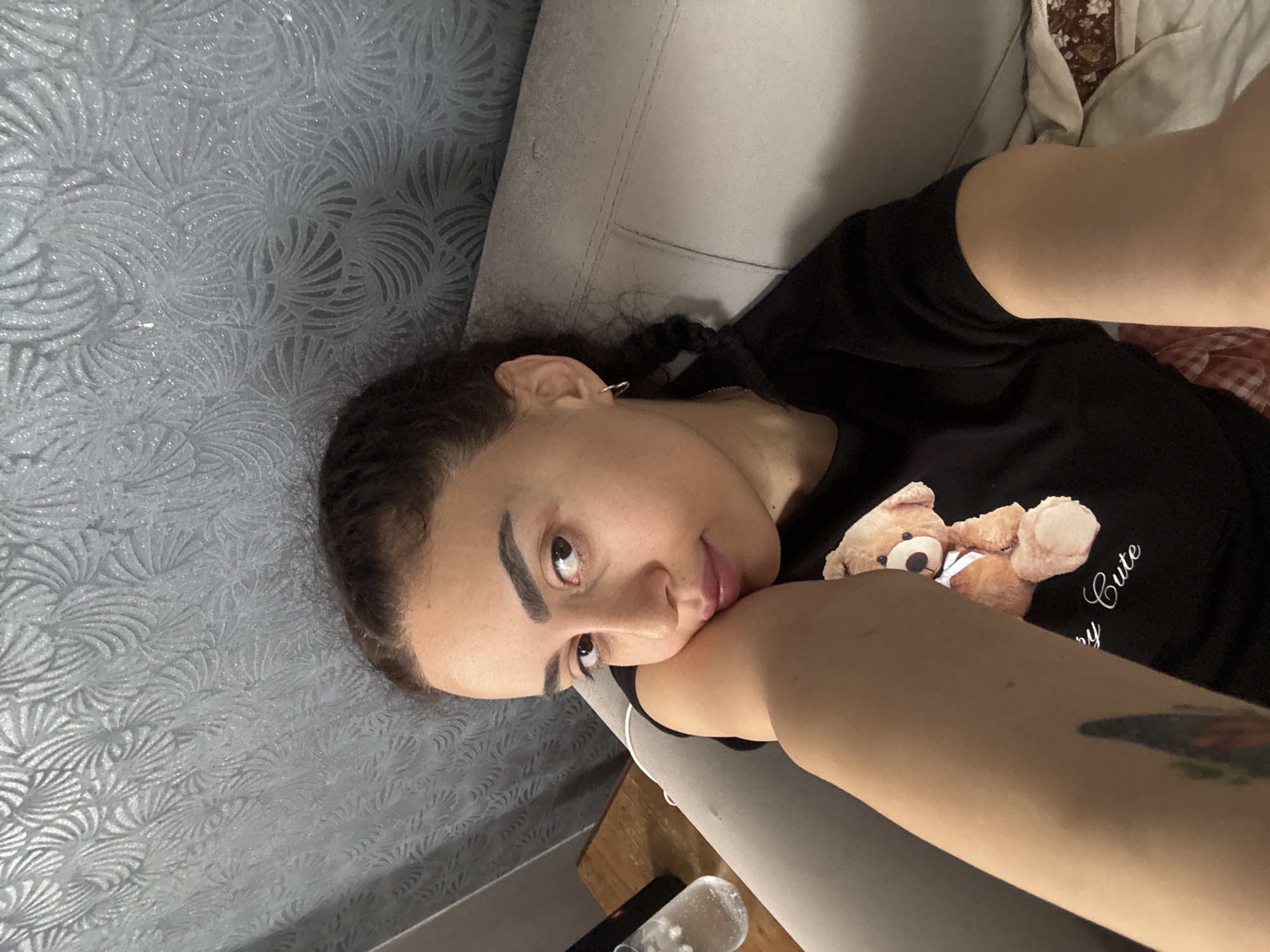 A woman with brown hair and dark eyebrows is laying on a gray sofa. She is wearing a black t-shirt with a cartoon bear on it. Her left arm is bent and she is resting her cheek on it. There is a glass bottle with white liquid inside it on the lower part of the image. The background is a gray wall with a floral pattern. I cannot specify the exact location within Tbilisi, Georgia based on this image.