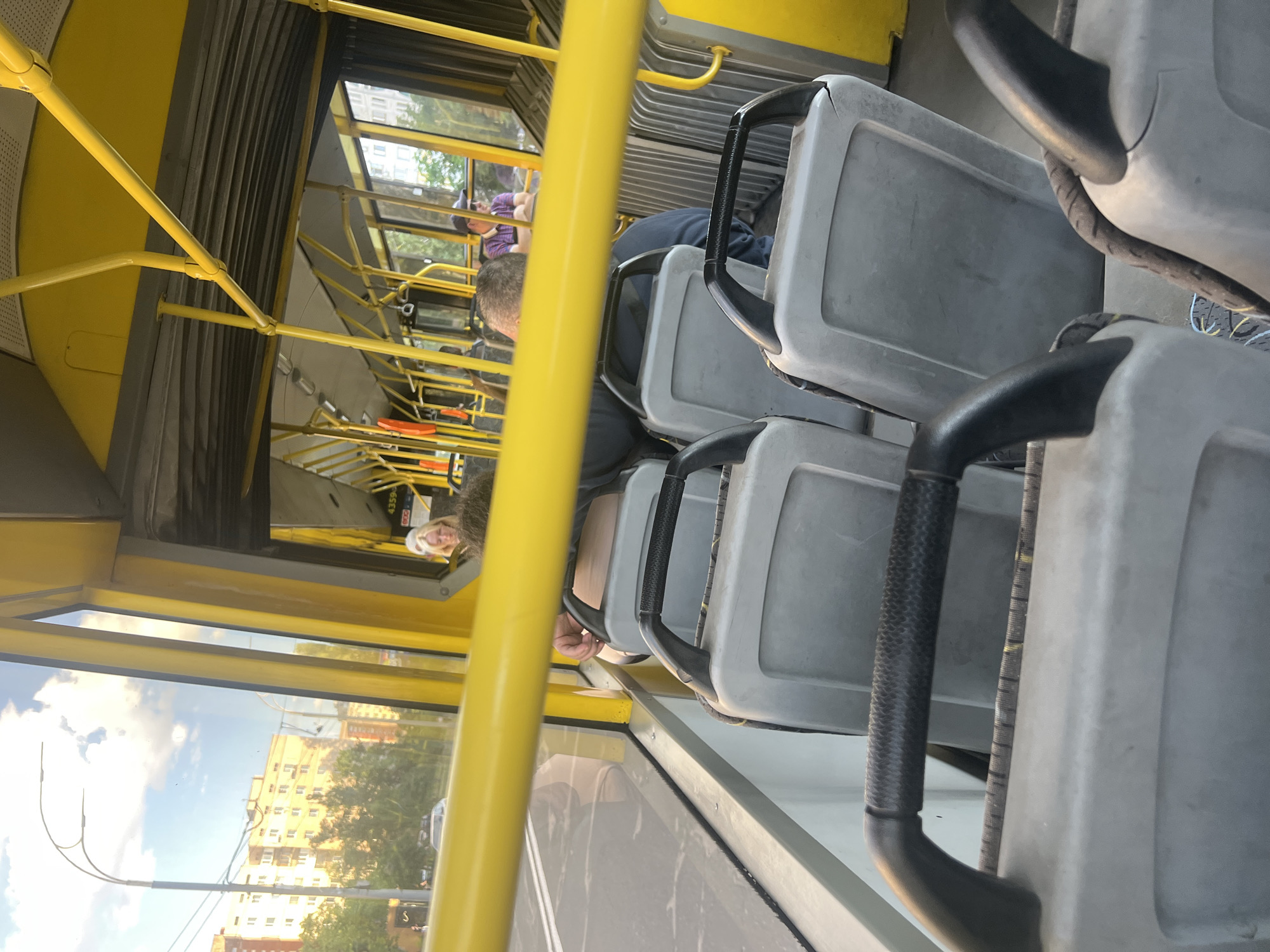 The image depicts the interior of a bus in Kyiv, Ukraine, with a view looking forward from a passenger's seat. The bus is yellow and gray, with yellow handrails and poles throughout. Several passengers are visible, including a woman with blonde hair in the front left of the bus, and a man wearing a hat and sunglasses further back on the right. Outside the window, a street scene with apartment buildings and trees is visible under a cloudy sky. The overall scene is consistent with public transportation within an urban environment.