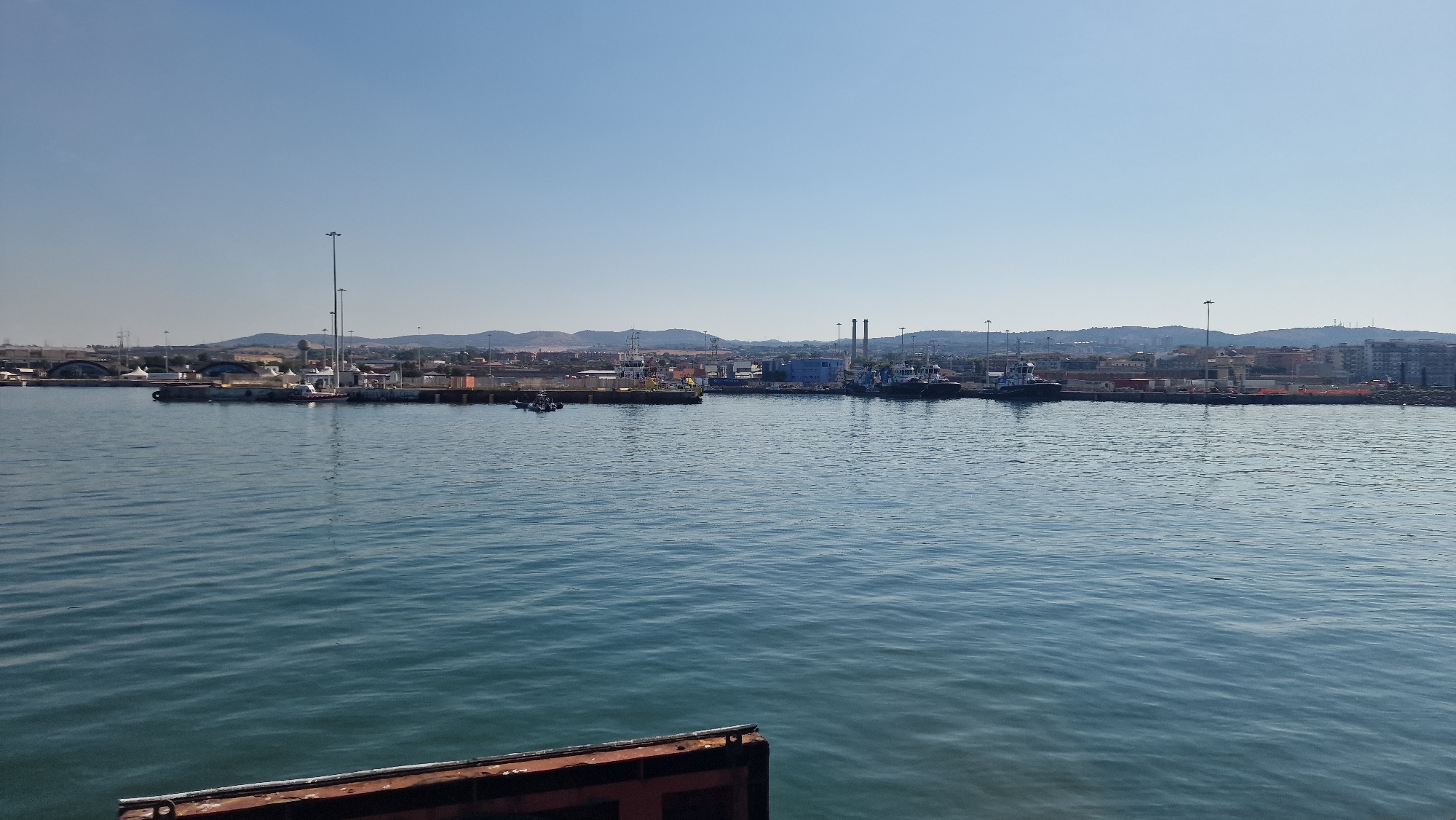 The image shows a wide view of the Civitavecchia, Italy harbor on a clear, sunny day. The foreground is dominated by the blue-green, rippling surface of the sea, with a small section of a dark, metallic structure visible at the bottom edge, likely part of a boat or dock.
In the mid-ground, the harbor is lined with various vessels. To the left, several tugboats are docked or anchored. Further into the harbor, a larger white vessel with a crane is prominent, along with other boats and industrial structures. The coastline beyond the harbor features a mix of industrial buildings, including what appear to be smokestacks, interspersed with urban development and residential buildings. Hills form the backdrop, with a clear, light blue sky above. The overall impression is of an active, working port with industrial and urban elements blending with the natural landscape.