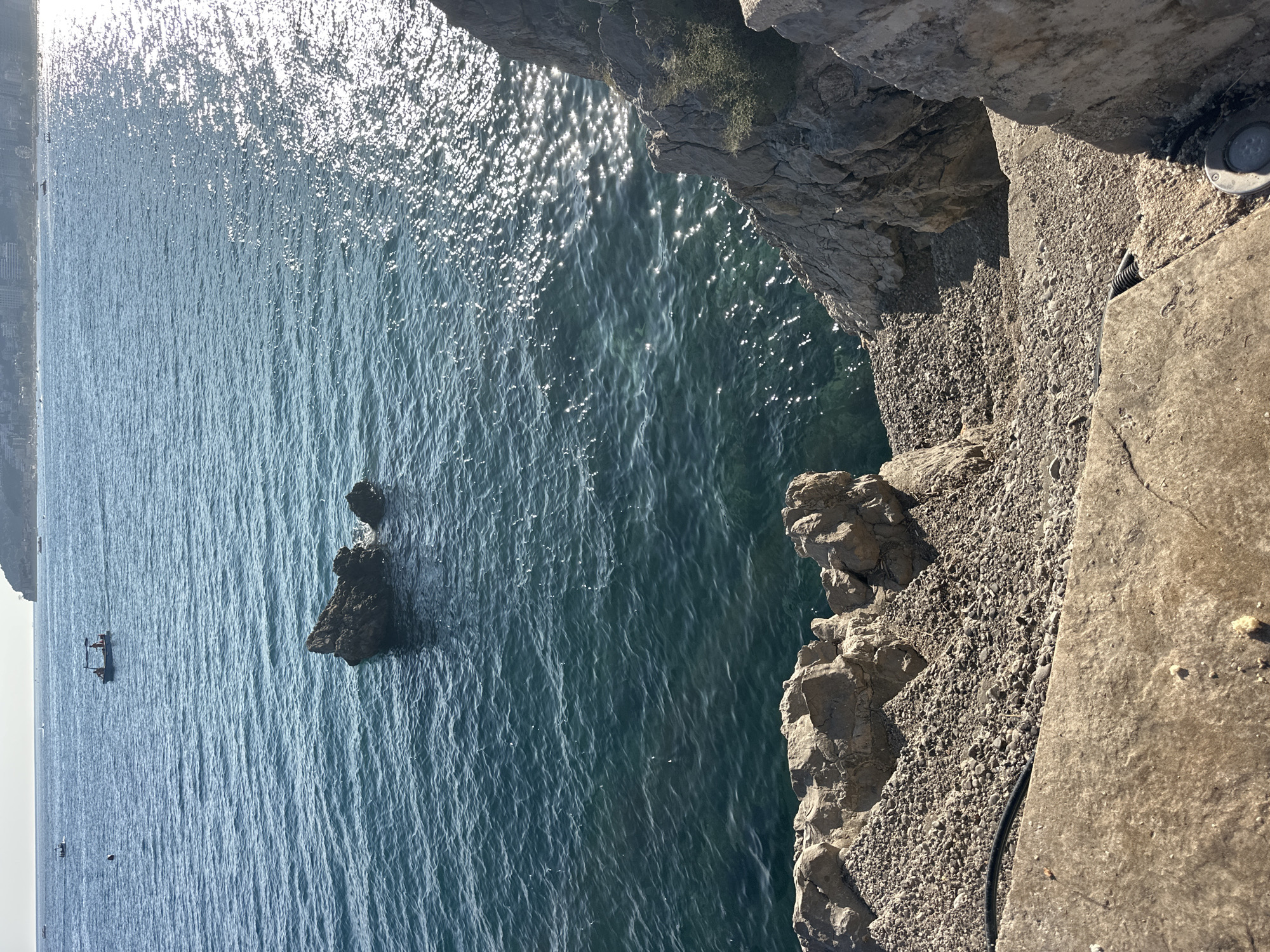 A high-angle shot shows a rocky coastline meeting the Adriatic Sea in Budva, Montenegro. The foreground features a concrete platform and a pathway made of gravel and stones leading down towards the water. Jagged, light-gray rocks line the right side of the frame, with sparse green vegetation growing from crevices. The sea is a deep turquoise, with sunlight creating shimmering reflections on its surface. Two dark rocks rise from the water in the mid-ground. In the distance, a faint line of buildings suggests the coastline of Budva, with a few small boats visible on the water. The sky is a hazy, light blue.