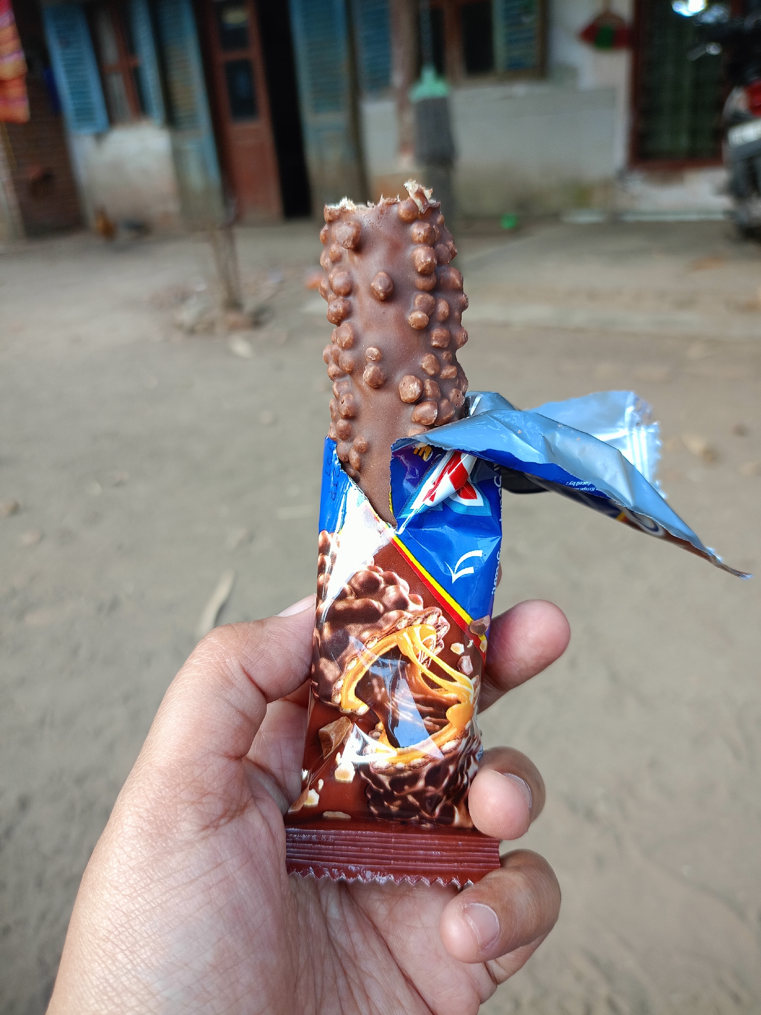 A hand holds a partially unwrapped chocolate bar, which has a rough, bumpy texture on its exterior. The chocolate bar is mostly covered by its blue and white wrapper, which has some red and yellow accents. The background is blurred but appears to be an outdoor scene with a sandy or dirt ground and some buildings in the distance. The image is taken from a slightly upward angle, with the hand and chocolate bar filling most of the foreground. The scene is likely in the city of Jelbuk, Indonesia.