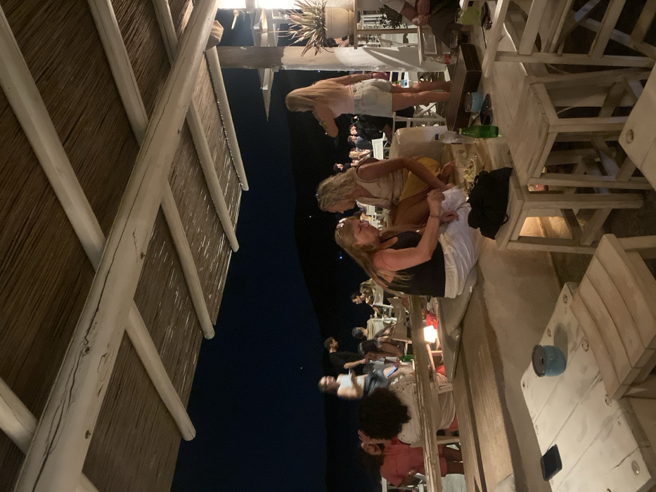 The image depicts an outdoor bar or restaurant scene at night in the city of Φάρος, Greece. Artificial lighting illuminates the immediate area, while the background reveals dark hills or landforms against a dark night sky.
The primary structure consists of a thatched roof supported by white-painted wooden beams and crossbeams. Below the roof, the establishment features white-painted, rustic-style wooden furniture. In the immediate foreground, a white wooden table holds a black smartphone and a light blue cylindrical container. Adjacent to this, a white wooden stool is visible. To the right, another white wooden table displays a green glass bottle, a dark object (possibly a small bag or case), and a rectangular wooden box. Two white wooden stools are also present near this table.
In the midground, a low, white-painted bench-like structure with white cushions runs horizontally. Two women are seated on this bench: one with blonde hair, wearing a black tank top and white trousers, is focused downwards, seemingly manipulating an object or food with her right hand. Beside her, another woman with blonde, curly hair, dressed in a light-colored sleeveless top and a yellow skirt, is seated facing her, observing. A small, glowing light source is positioned on the bench near them.
Further to the right, a woman with long blonde hair, wearing a light pink top and light-wash denim shorts, stands with her back partially to the viewer, her right arm raised, suggesting she is drinking from a glass. Near her, a potted plant with spiky green leaves rests on a white stand.
Throughout the midground and background, approximately 15-20 individuals are visible, seated and standing at various white-painted tables and chairs, engaged in social interactions. One individual in a dark shirt is seen standing and moving in the mid-distance. The overall scene suggests a relaxed, social gathering.
