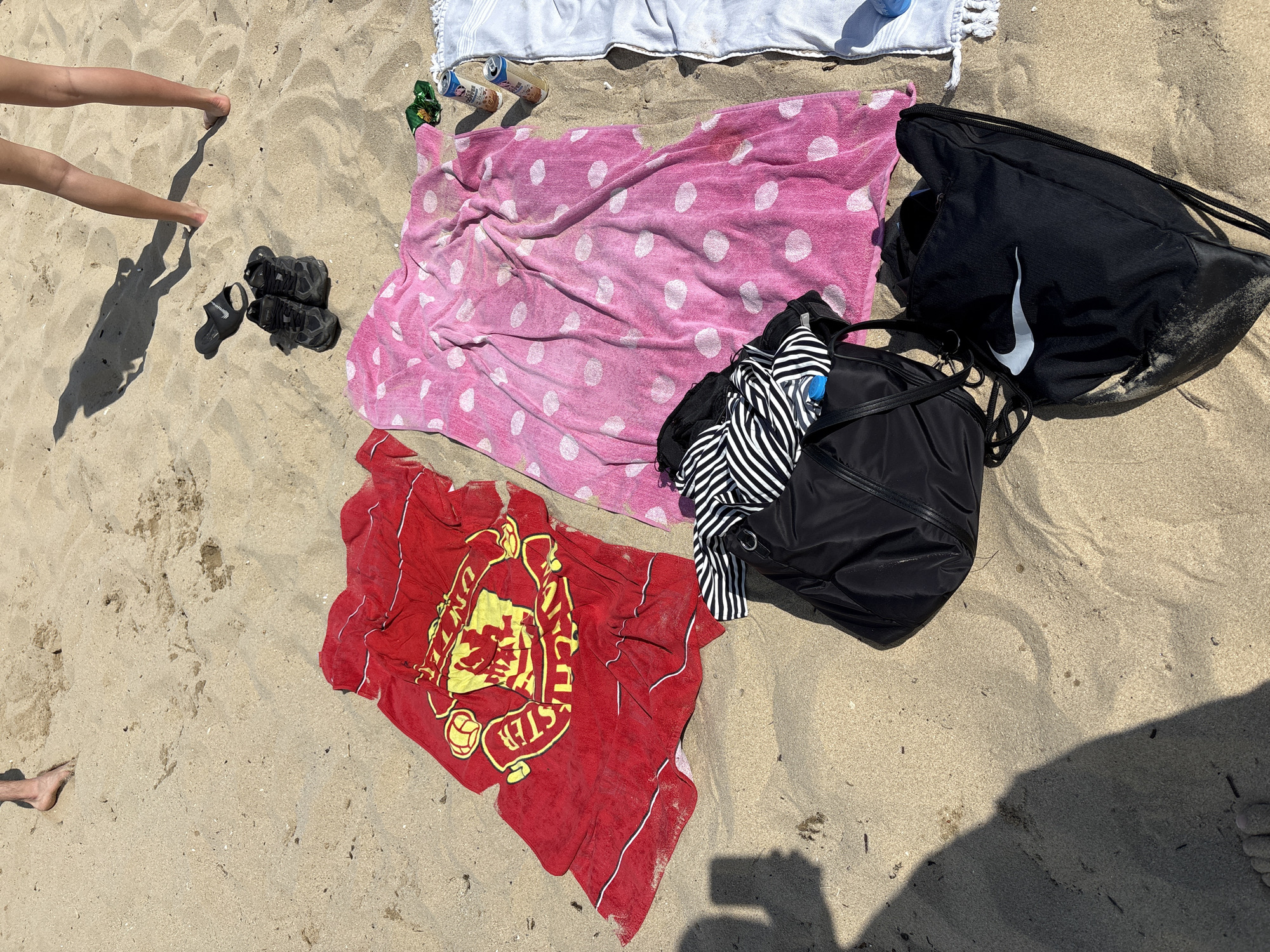 The image depicts a sunny day on a sandy beach, likely in Varna, Bulgaria, given the context. In the foreground, several items are laid out on the sand, suggesting a break or relaxation. A red towel with the Manchester United crest is prominent, next to a pink polka-dotted towel. Two black bags are visible: one partially open, revealing black and white striped clothing, and another, a Nike drawstring bag, lies flat. Nearby are a pair of black sneakers and a black slide sandal. Two cans of beverage are placed on a white towel in the upper right portion of the frame. In the background, the legs of at least two individuals are visible, standing or walking on the sand, casting long shadows. The environment is clearly a beach, with fine sand and scattered debris.