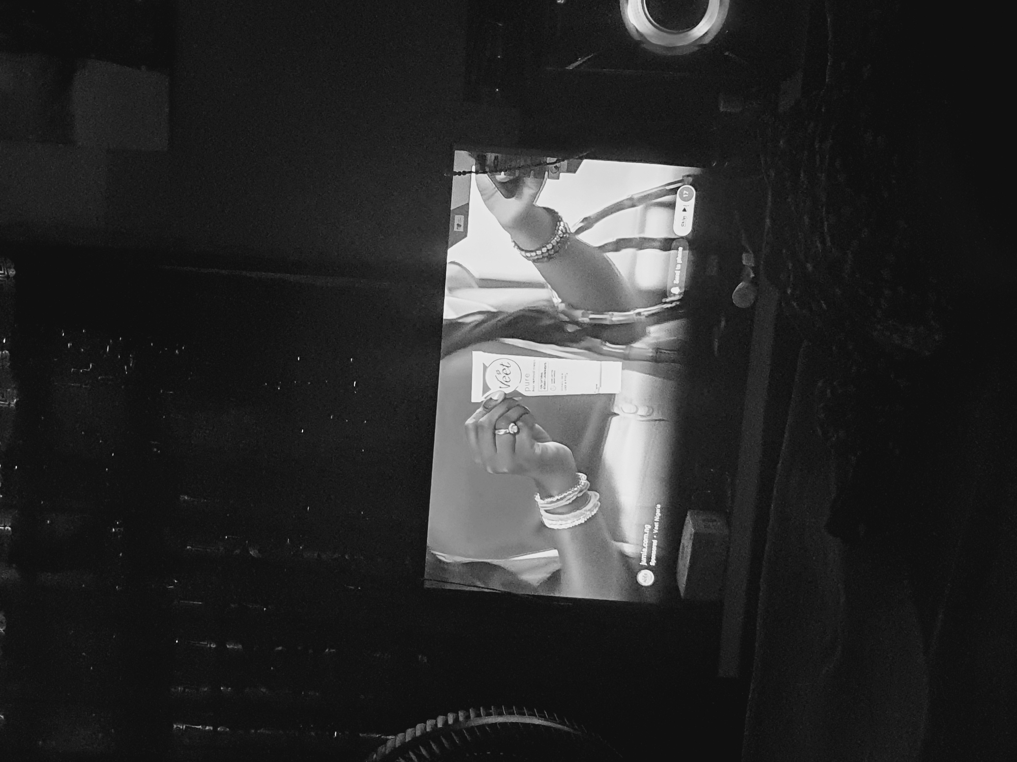 A black and white image displays a television screen showing an advertisement. A person's arm is featured in the ad, adorned with bracelets and a ring, holding a product labeled "Veet." The advertisement includes the text "jumia.com.ng Sponsored - Veet Nigeria." A "Skip" button with the number 17 is visible on the upper right corner of the screen, with the option to "Send to phone" underneath. The location is inferred as Lokoja, Nigeria, based on the product being advertised in Nigeria. The television is placed on a surface with some objects nearby, partially obscured by the darkness.