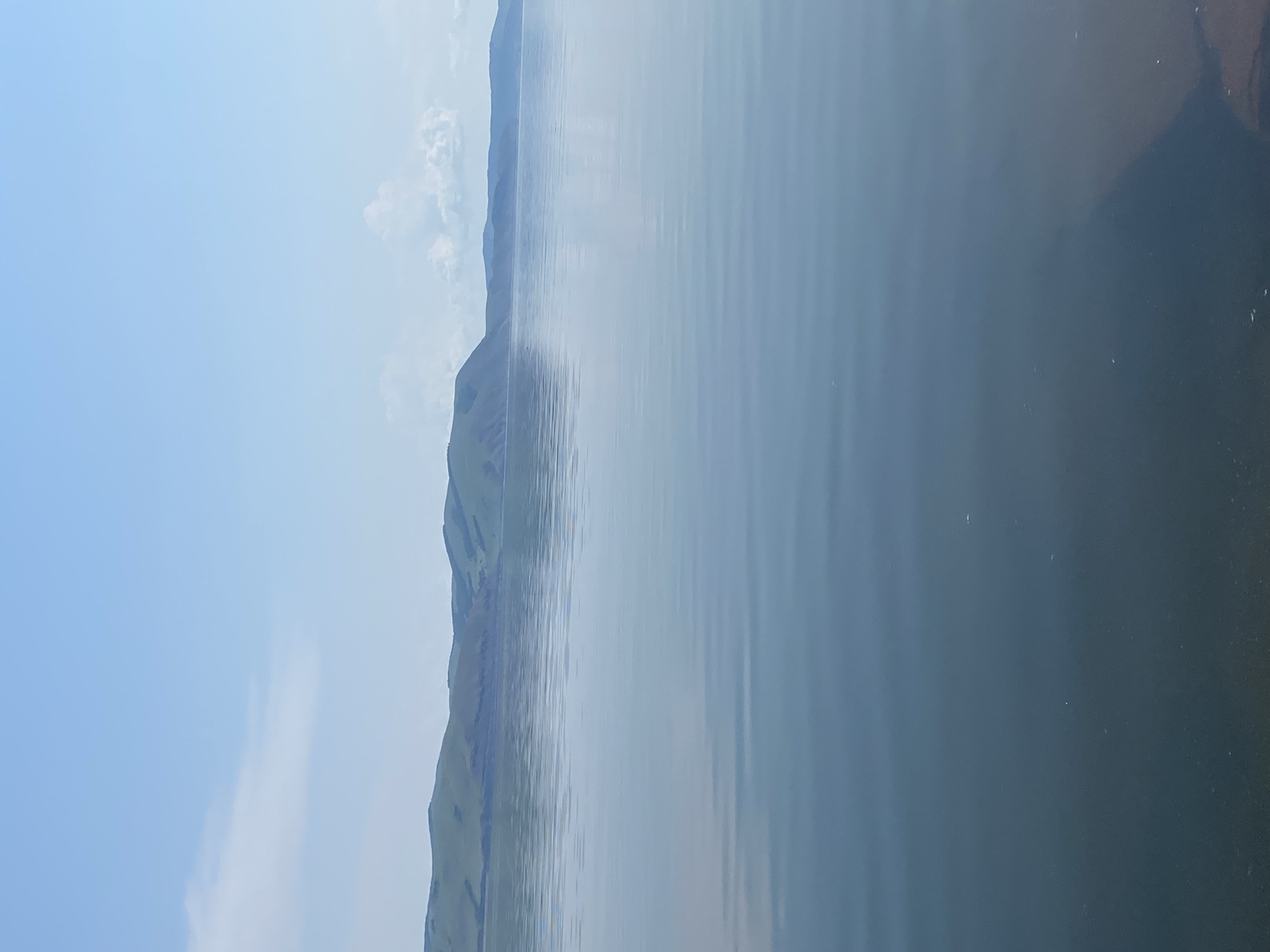 The image shows a serene landscape with a calm body of water in the foreground and rolling green hills in the background under a clear blue sky. The water exhibits gentle ripples, reflecting the soft light. The hills are covered in vegetation and appear to be part of a mountain range stretching across the horizon. Faint clouds are scattered across the sky, adding depth to the scene. The overall impression is one of natural tranquility. The location is identified as Ust'-Erba, Russia.