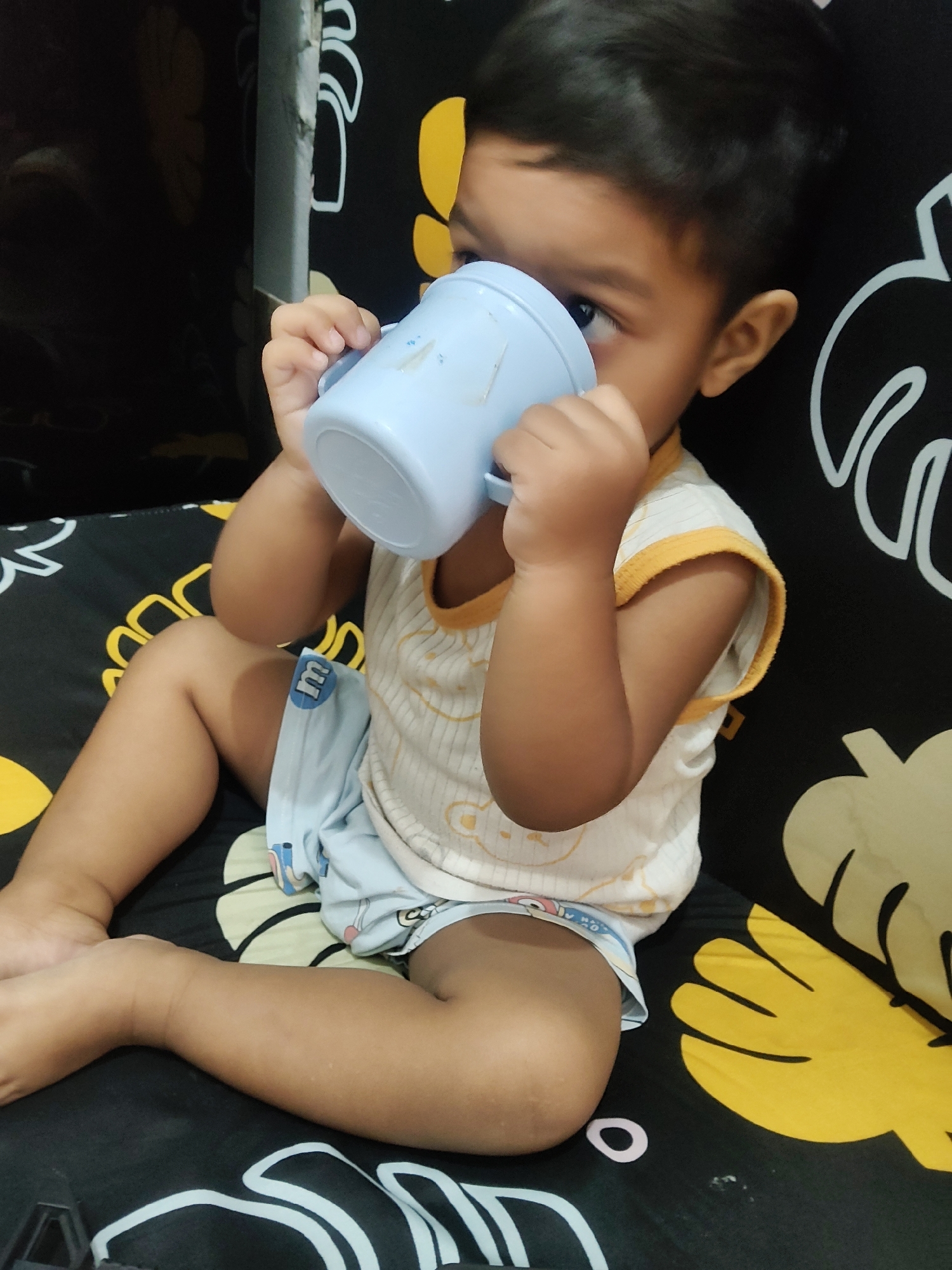 An upward angle shot shows a young child drinking from a light blue cup in Mekarmukti, Indonesia. The child is wearing a sleeveless white shirt with yellow trim and light blue shorts with an "M" logo. The child is sitting on a black surface with a repeating pattern of yellow flowers and white abstract shapes. The background is dark and out of focus.