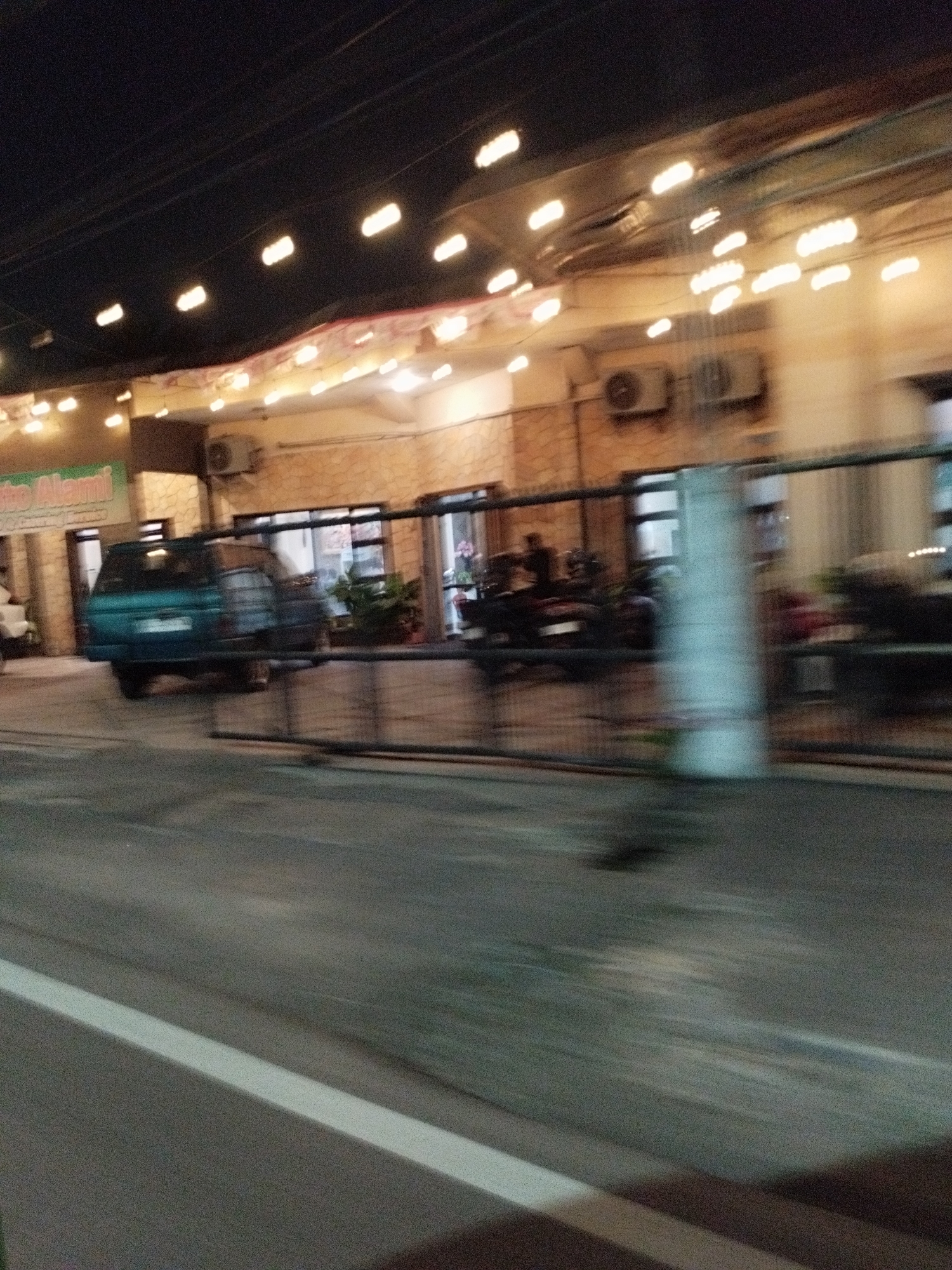 The image shows a blurred exterior view of a building at night, likely a commercial establishment, with a light-colored stone facade and a covered walkway with numerous overhead lights. A teal-colored van is parked in front of the building, to the left of a metal fence. Behind the fence, several motorcycles are visible. The ground in front of the building appears to be a paved area, and the street in the foreground has a white line marking. The image quality is poor due to motion blur and low light conditions. The context provided suggests this scene is in Surabaya, Indonesia.