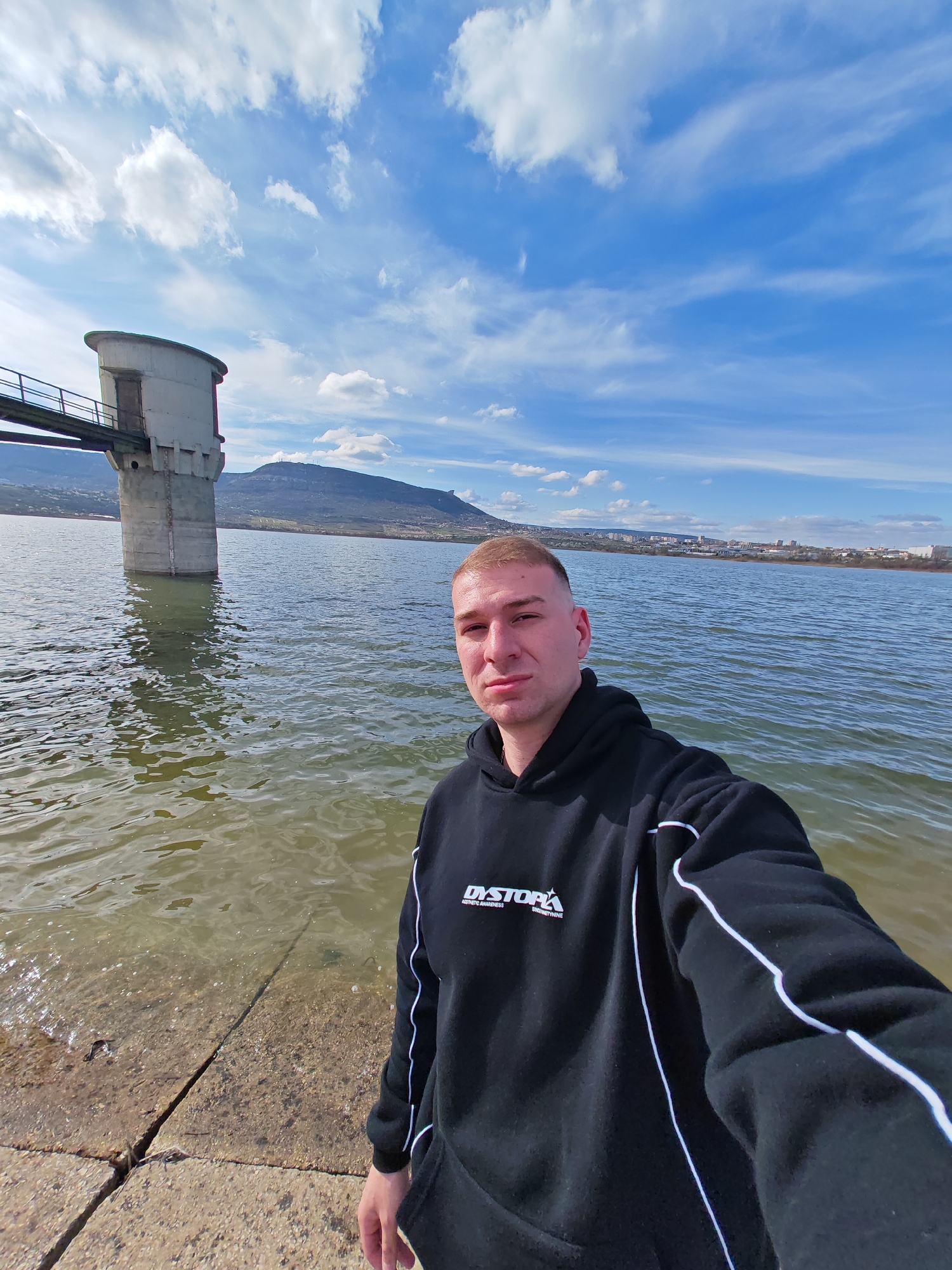 A man stands at the edge of a body of water, near a concrete structure connected to a tower in the water by a bridge. The man is wearing a black hoodie with white stripes on the sleeves and a white logo on the chest. Behind him is the lake of Kyosha in the city of Shumen, Bulgaria, with a distant mountain in the background. The sky is blue with scattered clouds. The water is choppy.