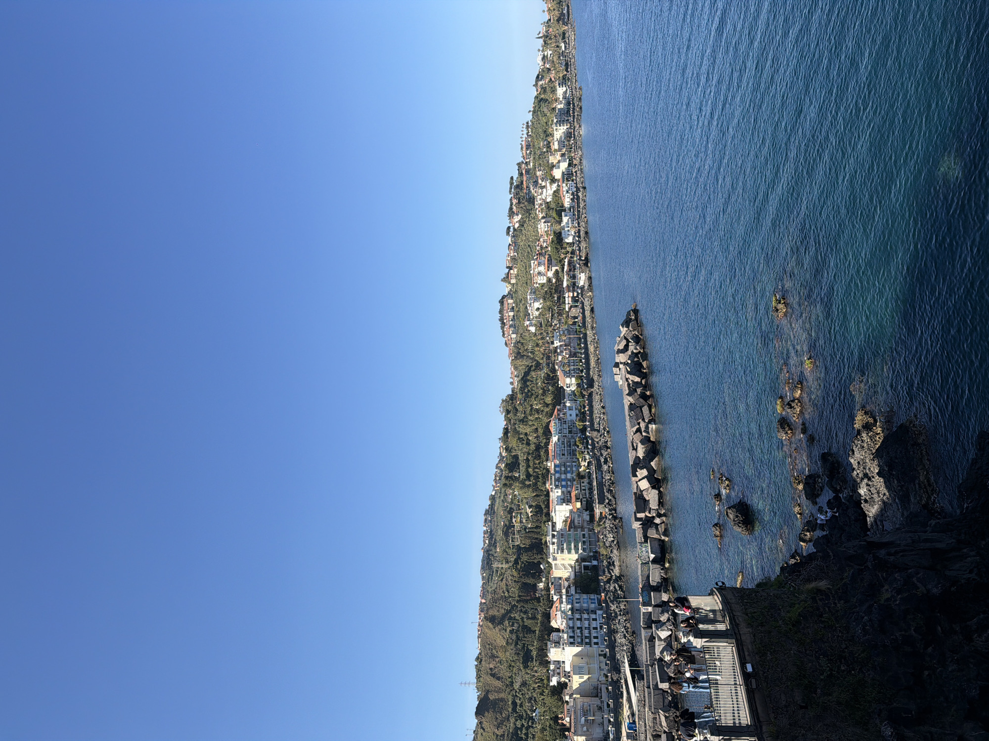 The image shows a coastal view in Aci Castello, Italy. The scene is divided between a clear blue sky and a landscape of land and sea. Along the coastline are buildings and trees, with a small town situated on the hillside. A breakwater of large stone blocks extends into the sea. People are present on a platform near the breakwater. The sea is blue with visible rocks near the coast.