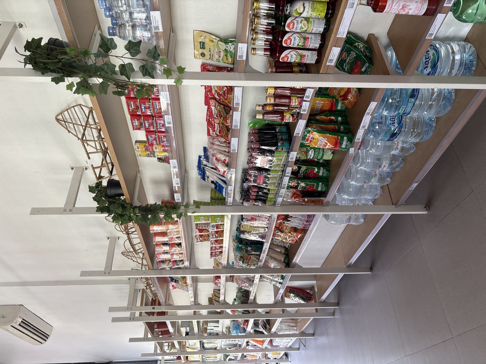 The image depicts the interior of a small grocery store, likely a minimarket, located in Gajahan, Indonesia. The store features multiple shelves stocked with various food items, drinks, and household goods. On the far left, an air conditioning unit is mounted high on the wall. The shelves are made of metal frames with wooden planks.
The shelves on the left are filled with items such as canned goods, packaged snacks, and bottled drinks. Further to the right, the shelves display a variety of packaged foods, including instant noodles, seasonings, sauces, and rice. There are also several bottles of water, including brands like "Le Minerale" and "Aqua," prominently displayed on the lower shelves. Decorative artificial vines are draped from the upper shelves on the right side of the image, adding a touch of greenery to the store's interior.
The overall scene is well-lit and organized, suggesting a typical retail environment. No people are visible in the image. The flooring is tiled with a neutral gray color. The store appears to be a local establishment catering to everyday needs.