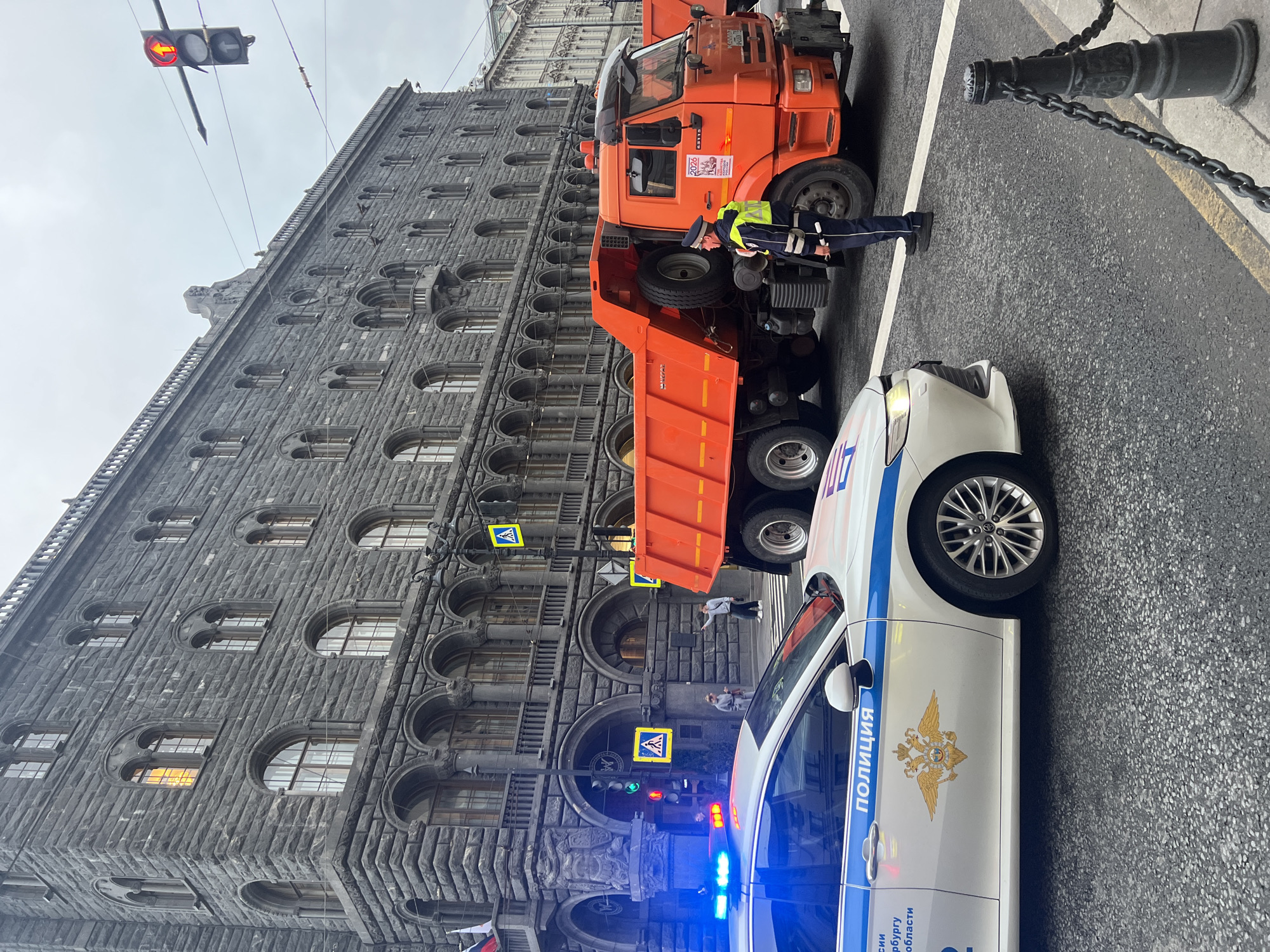 The image depicts a street scene in Saint Petersburg, Russia, with a police officer interacting with the driver of a large orange dump truck. The truck's cargo bed is raised, suggesting it may have been involved in some kind of incident. A white police car with blue flashing lights is parked nearby. The building in the background is a large, grey stone structure with arched windows, indicating a historical or government building. Traffic lights are visible, with one showing a red arrow. The scene appears to be taking place on a city street, with pedestrian crossing signs also present.