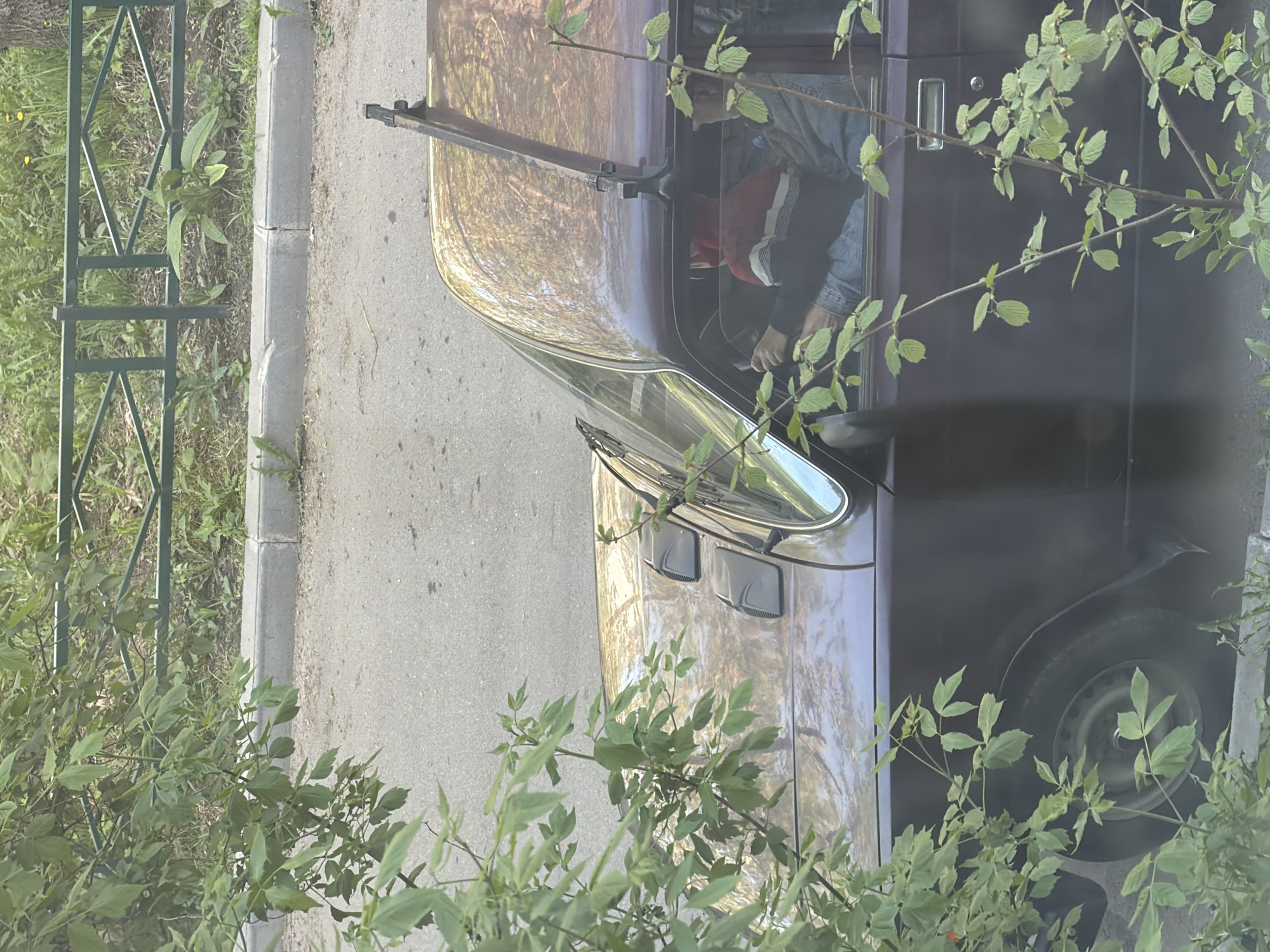 The image depicts an older, dark-colored car parked next to a curb in Tokkari-Lend, Russia. A person is visible inside the car. The car is situated next to a sidewalk, with vegetation, including leafy branches, partially obscuring the view. A green metal fence is visible to the left.