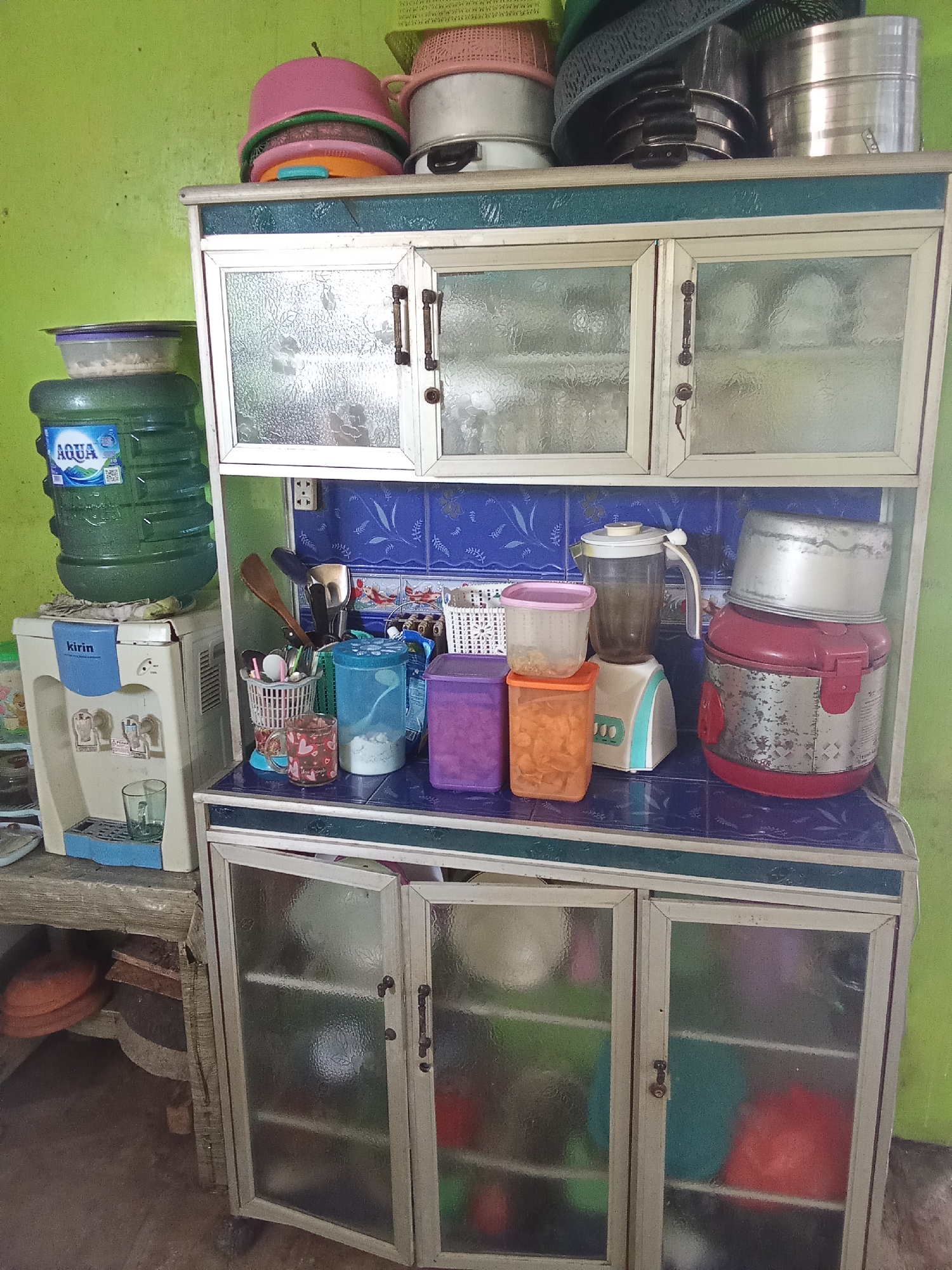 An indoor scene shows a cluttered kitchen cupboard filled with various containers, appliances, and cooking utensils. The cupboard is made of aluminum and glass, with a turquoise trim. On top of the cupboard, there are several metal pots and pans, some plastic bowls, and a woven mat.
On the shelf of the cupboard, there is a water dispenser with a large blue Aqua water bottle on top. Next to it, there is a blender, several plastic food containers, and a pink rice cooker with a metal pot on top. There is also a collection of cutlery in a basket, a blue pitcher, and a mug with a heart pattern.
The bottom section of the cupboard has glass doors, revealing various colorful containers inside, including a red one, a green one, and a blue one. The cupboard is placed against a lime green wall. A portion of a wooden shelf with some round orange objects is visible to the left of the cupboard.
The image appears to have been taken in a residential kitchen, possibly in Subang, Indonesia, given the presence of an Aqua water bottle, a popular brand in Indonesia, and the general aesthetic of the kitchen.