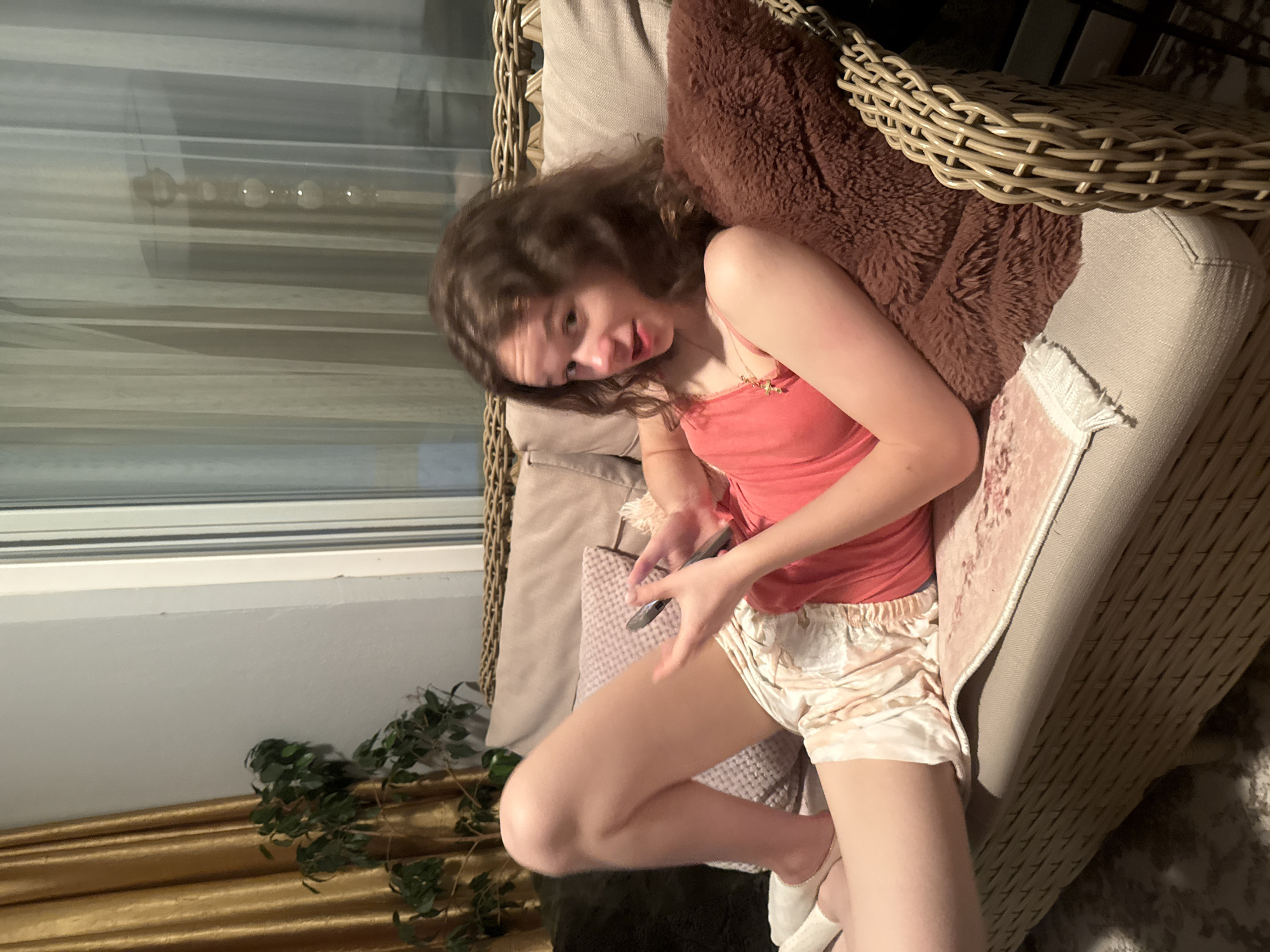 A young woman sits in a wicker chair, holding a mobile phone. She wears a coral-colored tank top and light, satin shorts. She is leaning back against a brown, furry pillow. The chair is placed near a window with closed blinds and a curtain. There is a potted plant nearby. The location is Unknown, Unknown.