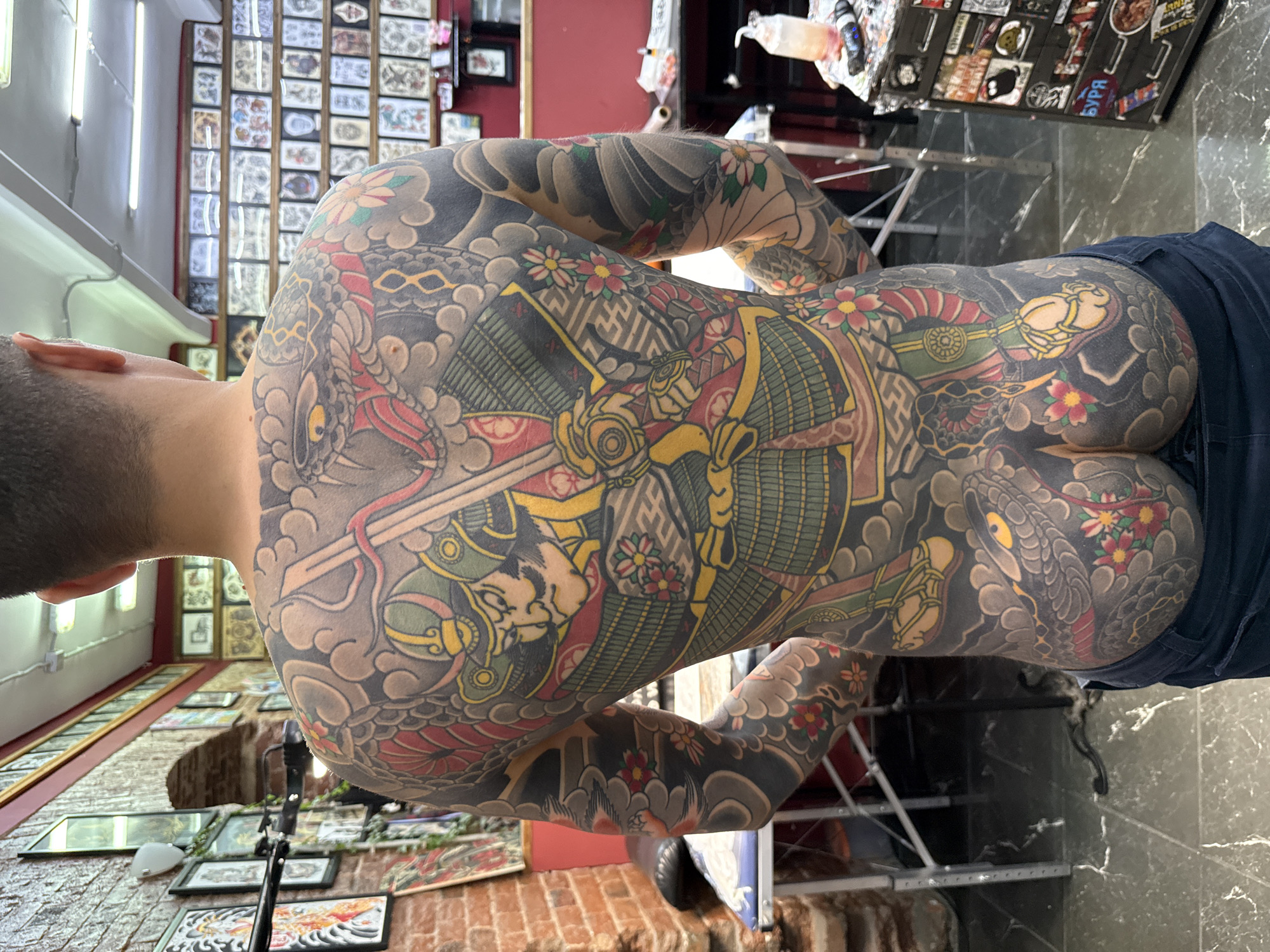 A man is pictured from the back, displaying extensive full-body tattoos. The tattoos appear to depict imagery commonly associated with Japanese artistic themes, including samurai, dragons, and flowers. The man is wearing what appears to be denim pants. The backdrop reveals what looks like a tattoo parlor, with walls adorned with tattoo designs. In the background, a sign displays "БУРЯ", which suggests the city of Moscow, Russia. The image seems to be taken indoors in a well-lit environment, possibly within the tattoo establishment.
