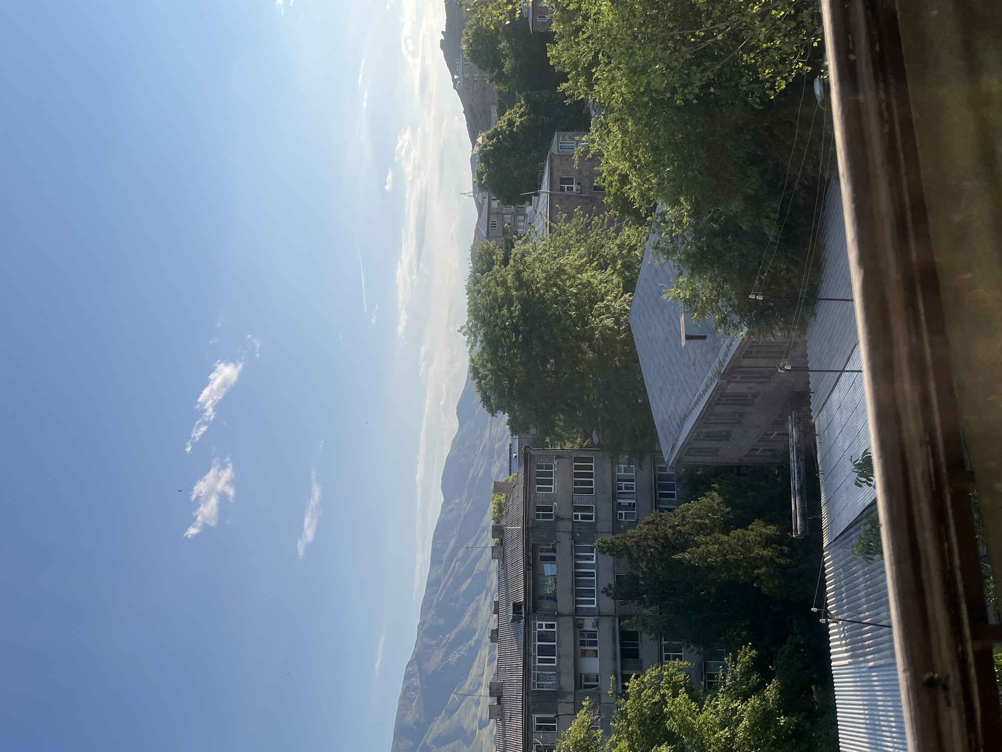 The image captures a view of urban buildings and natural landscape in Gladzor, Armenia, under a clear blue sky with scattered white clouds. In the foreground, a wooden railing suggests the viewpoint is from a balcony or window.
The middle ground is dominated by several residential buildings. One prominent building on the left is a multi-story concrete structure with numerous windows, its roof covered in grey corrugated material. Adjacent to it are tall, dark green coniferous trees. To the right, other apartment buildings are visible, some partially obscured by dense green foliage from deciduous trees. One building in the mid-right has a grey, pitched roof.
The background features a range of layered mountains, characterized by their undulating slopes and arid, rocky appearance, indicative of a continental climate. The sunlight casts shadows and highlights, suggesting it is either morning or late afternoon. There are no people visible in the scene. The overall atmosphere is peaceful and somewhat rustic, with the urban elements integrated into a natural setting.