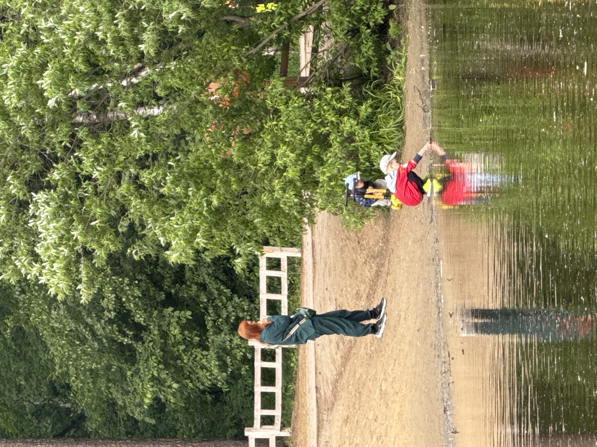 A horizontal outdoor scene depicts a section of a wooded area adjacent to a body of water, possibly a river or lake. On the right, the water reflects the sky and foliage, showing slight ripples and disturbances on the surface.
A pathway runs parallel to the water's edge, where a small child is sitting, dressed in a red and black outfit with a white cap, and appears to be reaching towards the water. Near the child, there's a collection of bags and equipment, possibly fishing or camping gear.
On the left, a person with reddish hair, clad in a green jumpsuit, is standing on the pathway near a white wooden fence-like structure. This person appears to be looking towards the water.
The background is filled with dense green trees, indicating a natural setting. The scene seems to be taken on a sunny day, judging by the reflections and shadows.
Without additional information, a specific location in Perm, Russia cannot be determined.