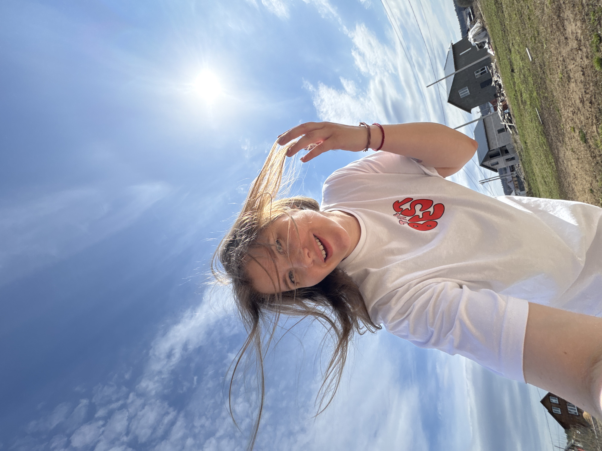 An outdoor, low-angle selfie captures a young woman smiling and looking down at the camera, against a bright blue sky with scattered clouds. The image is taken in Gornyi (Dvurechenskoe S/P), Russia. The woman has long, light brown hair, with some strands wind-blown across her face. She wears a white long-sleeved t-shirt featuring a stylized red graphic, possibly resembling a lobster or abstract shape, on the front. On her right wrist, she has a red beaded bracelet and another string-like bracelet. Her left hand is raised, touching her hair near her head.
The bright sun, positioned high in the sky to the upper left, indicates it is daytime, likely late morning or afternoon. The weather is clear and sunny. In the background, visible to the right and appearing distant, are residential structures. One house is dark grey with white-framed windows, and another appears to be a lighter, possibly concrete or unfinished building. Utility poles or power lines are also discernible in the background. The ground beneath the buildings appears to be an expanse of bare earth and sparse grass, suggesting a rural or developing area. The woman's expression is pleasant and relaxed, with a slight smile.