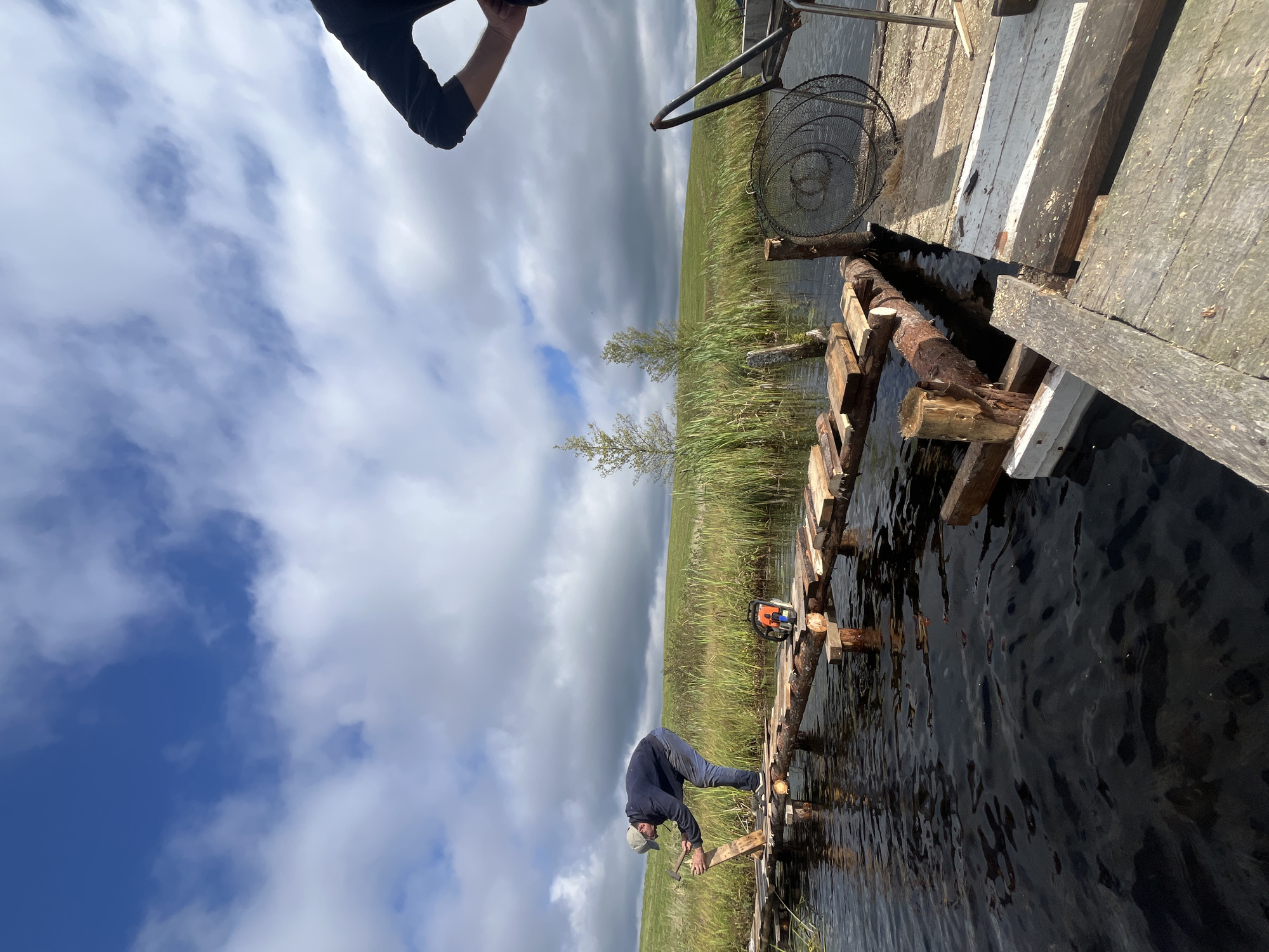 The image depicts a rural outdoor scene in the Дунілавіцкі сельскі Савет, Belarus. In the foreground, a man wearing a cap, dark jacket, and jeans is actively constructing a makeshift wooden bridge over a body of dark, rippling water. He is holding a hammer in his right hand and appears to be securing a wooden plank onto the partially built structure. A chainsaw rests on the bridge near him. The bridge is constructed from rough-hewn logs and planks, supported by vertical wooden posts. To the right of the bridge, a wooden platform is visible, with a black wire fishing net and some debris. In the background, a grassy, rolling hillside stretches under a sky with scattered white clouds. Two small trees are visible on the left side of the background. Another person, wearing a black long-sleeved shirt, stands on the wooden platform on the right, with their back partially to the camera. The overall impression is of a construction project in a natural, somewhat unrefined setting.