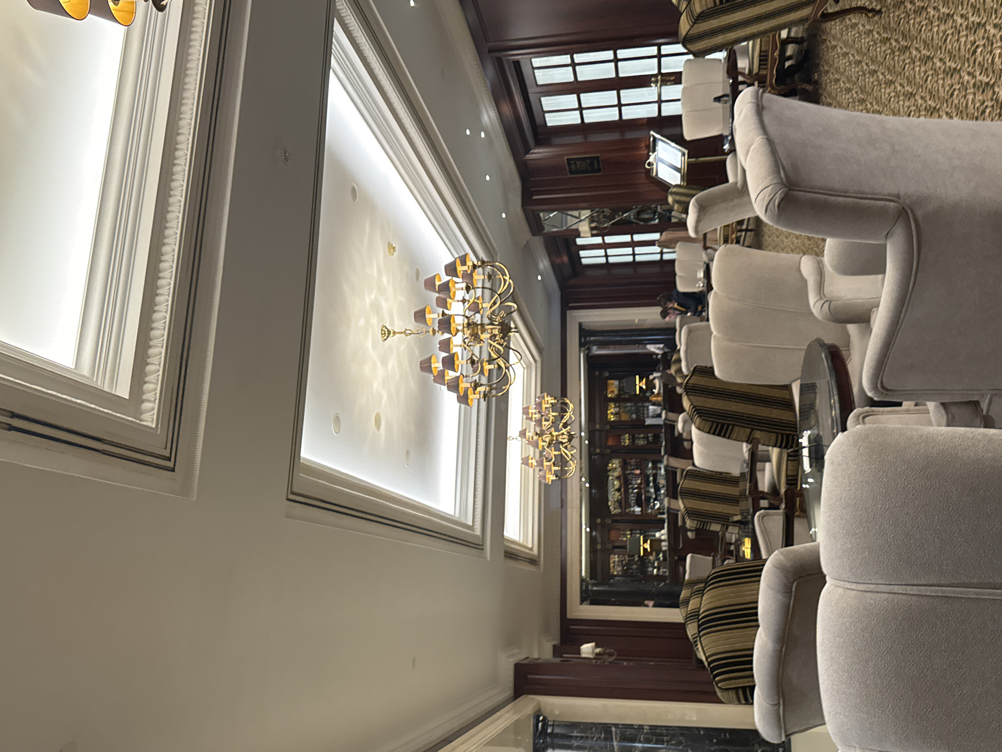 The image depicts the interior of what appears to be a high-end bar or lounge in Kyiv, Ukraine. The setting is characterized by ornate architectural details, including decorative molding on the walls, and ornate chandeliers. The seating area features high-backed, light-colored armchairs arranged near small tables, complemented by striped upholstered chairs. A bar area is visible in the background. Patrons are seen at the bar. The environment exudes a sense of luxury and sophistication.