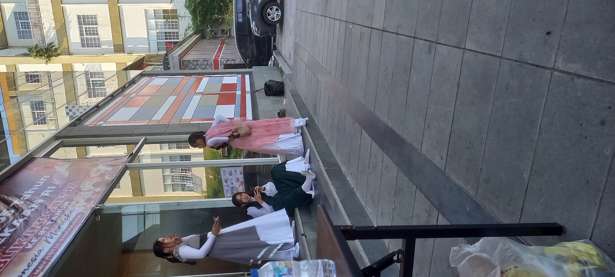 Three young women are gathered on the steps of a building with a glass facade in Samarinda, Indonesia. The woman on the left is standing, wearing a grey and white dress, and holding a smartphone. The woman in the center is seated on the steps, also wearing a green dress, and is looking at her smartphone. The woman on the right is standing, wearing a pink and white outfit, with a small purse slung over her shoulder. A banner with Indonesian text is visible above the entrance. The building's exterior features a geometric pattern of colored rectangles and numerous windows. Paved walkways and two parked cars, one black and one grey, are visible in the background. A black trash bag and a plastic bag with yellow contents are on the ground near the steps. The scene appears to be outdoors during daylight, with the sun casting shadows.