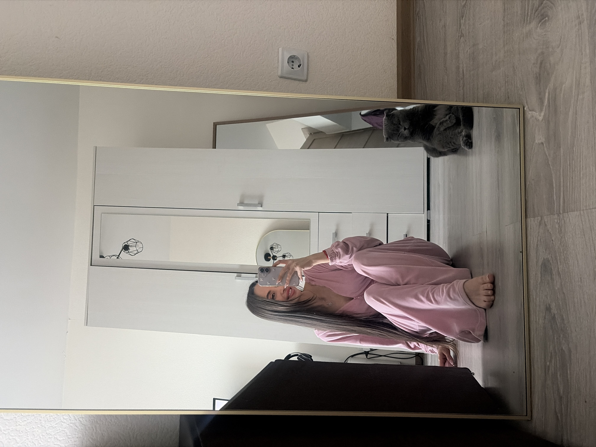 A mirror reflection shows a young woman in pink loungewear taking a selfie with a smartphone in what appears to be an indoor setting. In the reflection, a grey cat is visible on the wooden floor near a white cabinet. A framed mirror hangs above the cabinet. An electrical outlet is on the wall above the framed mirror. A dark-colored object, possibly a television or a piece of furniture, is in the foreground near the bottom of the mirror. The location cannot be determined from the provided image.