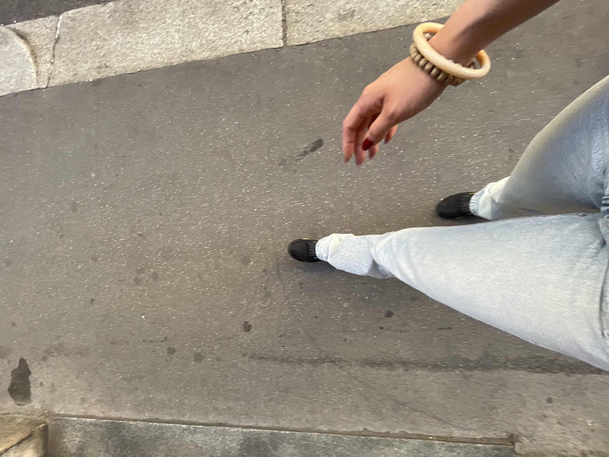 The image depicts a person standing on a grey pavement, likely a sidewalk or pedestrian area. The person is wearing light blue jeans, black shoes, and a beaded bracelet. The person's right hand is visible, with red nail polish on the fingernails. The pavement shows minor discoloration and stains. The lower left corner shows the edge of a concrete structure, possibly a curb or building foundation. There is no explicit confirmation of the location being Milan, Italy, in the given image.