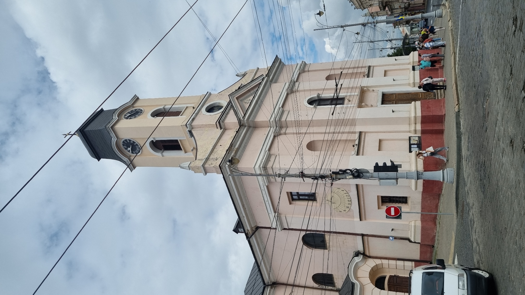 The image shows a pale pink church with a tall clock tower against a cloudy sky. A white van is parked to the left, and several people are walking on the cobblestone street in front of the church. There are numerous overhead wires crossing the frame. The clock tower has two visible faces with Roman numerals. A sundial is mounted on the side of the church. A red and white "no entry" sign is visible near the bottom left. The architecture suggests a European city, and the Ukrainian flag can be seen on a pole in the distance on the right. This is the Church of the Holy Spirit in Chernivtsi, Ukraine, a prominent landmark located at the intersection of Theater Square and Kobylianska Street.