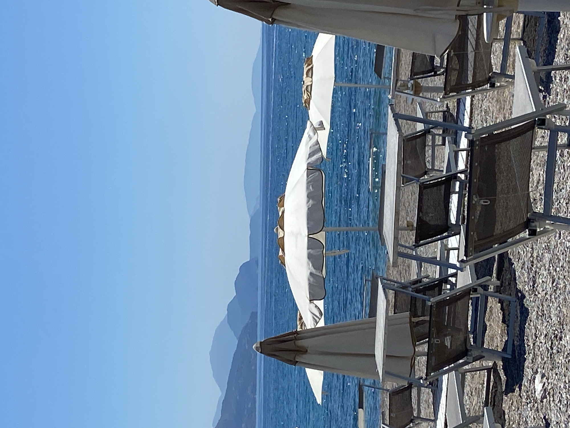 The image shows a beach scene in Prijevor, Montenegro. Several white parasols are positioned on the beach, with lounge chairs arranged underneath them. The beach appears to be composed of small pebbles. The sea is visible in the background, along with distant mountains. The sky is clear and blue. No people are visible in the scene.