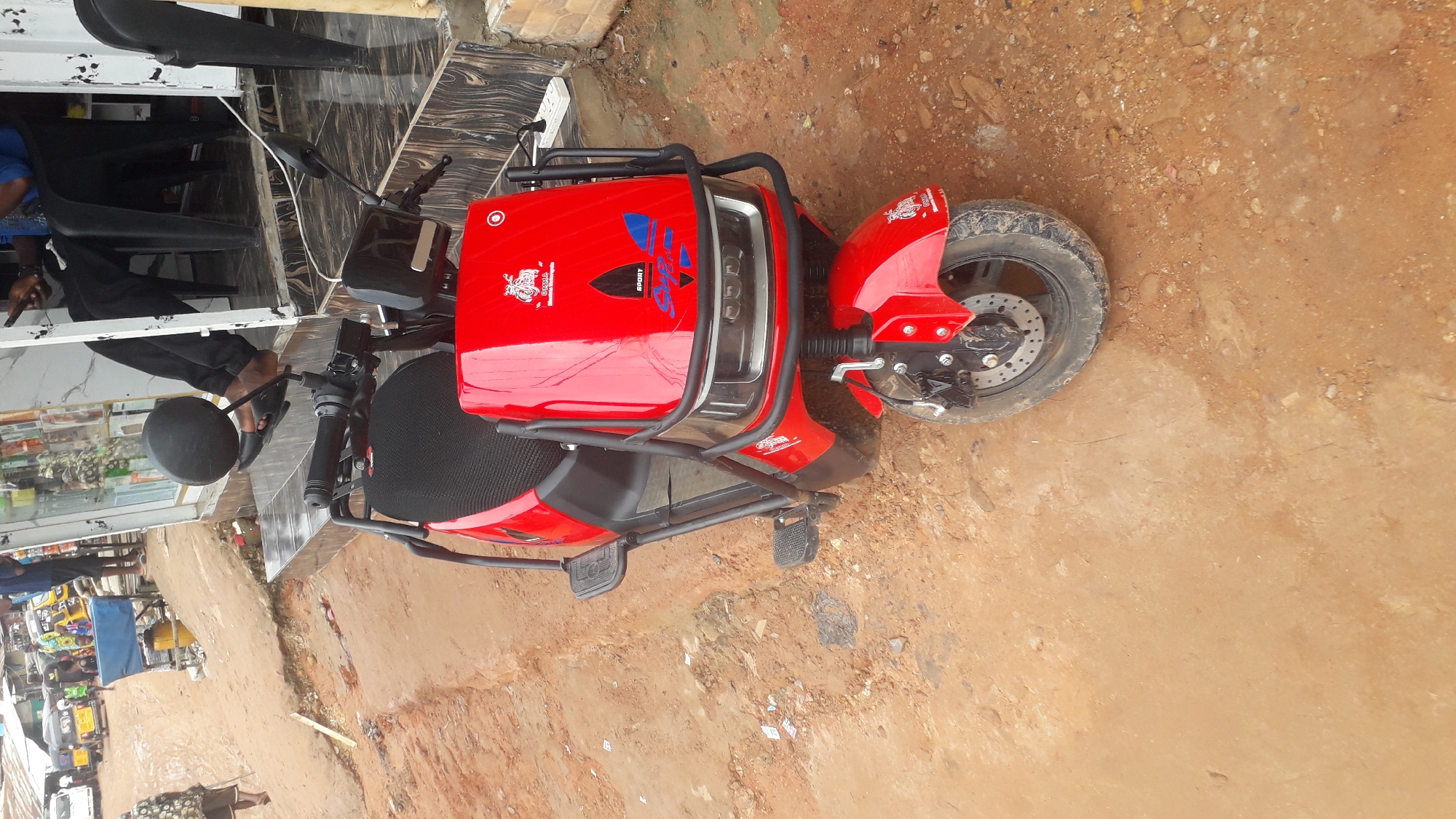 A red electric scooter is parked on a dirt ground in Nkpor, Nigeria. The scooter has black handlebars, a black seat, and black frame components. A person wearing black pants and sandals is partially visible near a building with a tiled facade. Other people and objects are blurred in the background.