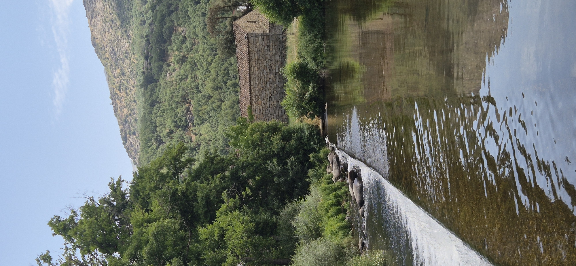 The image depicts a serene landscape scene. In the foreground, a gentle waterfall cascades over a series of large, dark rocks into a wide, calm river. The water's surface is slightly rippled, reflecting the surrounding greenery. The riverbank is lined with lush vegetation, including trees and bushes, with some white flowers visible amongst them.
To the right of the river, a rustic stone building with a terracotta tiled roof stands nestled amongst the trees. The building appears aged, constructed from irregular stone blocks. Beyond the building, a densely forested hillside rises, covered in a variety of green trees. The top of the hill is rocky and arid, with sparse vegetation. The sky above is a clear, pale blue. The overall impression is one of a tranquil, natural setting, likely in a rural or mountainous area.
