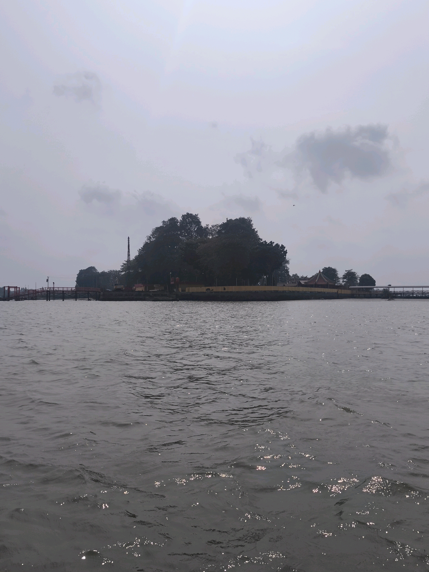 The image shows a wide body of water with a distant island in the center. The island is covered with dense trees and features several structures, including a prominent red-roofed building with ornate eaves and a tall, slender spire-like monument. A long, red-railed bridge extends from the left side of the image towards the island, and another bridge, appearing to be wooden and covered, is visible on the right. The water is choppy, with small waves reflecting the overcast sky. The sky is filled with diffused, light gray clouds. The scene appears to be the viewpoint from a boat on the Musi River in Palembang, Indonesia, looking towards Pulau Kemaro, an island known for its Chinese temple and spiritual significance.
