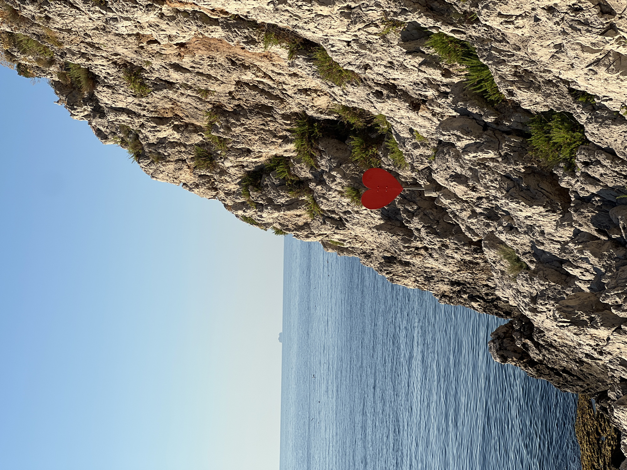 A bright red heart-shaped sign is affixed to the rocky cliff face overlooking the sea in Santa Flavia, Italy. The sign is positioned at mid-height on the cliff, and appears to be made of metal. The cliff is rugged and uneven, with sparse green vegetation growing in its crevices. The sea is a deep blue, with small waves and a distant ship visible on the horizon. The sky is clear and blue. The scene is bathed in the warm light of what appears to be either sunrise or sunset.